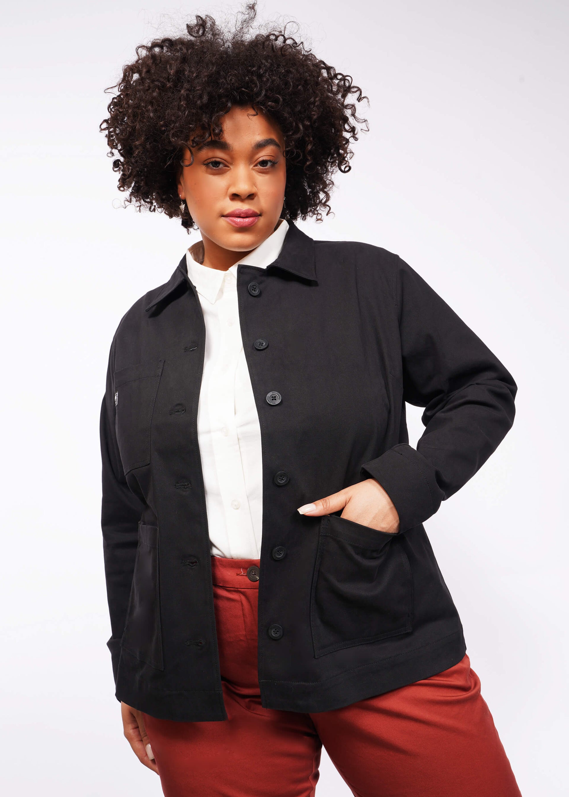 A person with curly hair models The Essential Utility Jacket, a sleek black utility jacket over a white shirt and red pants, standing against a plain white background. It features deep pockets that offer practicality without compromising style.