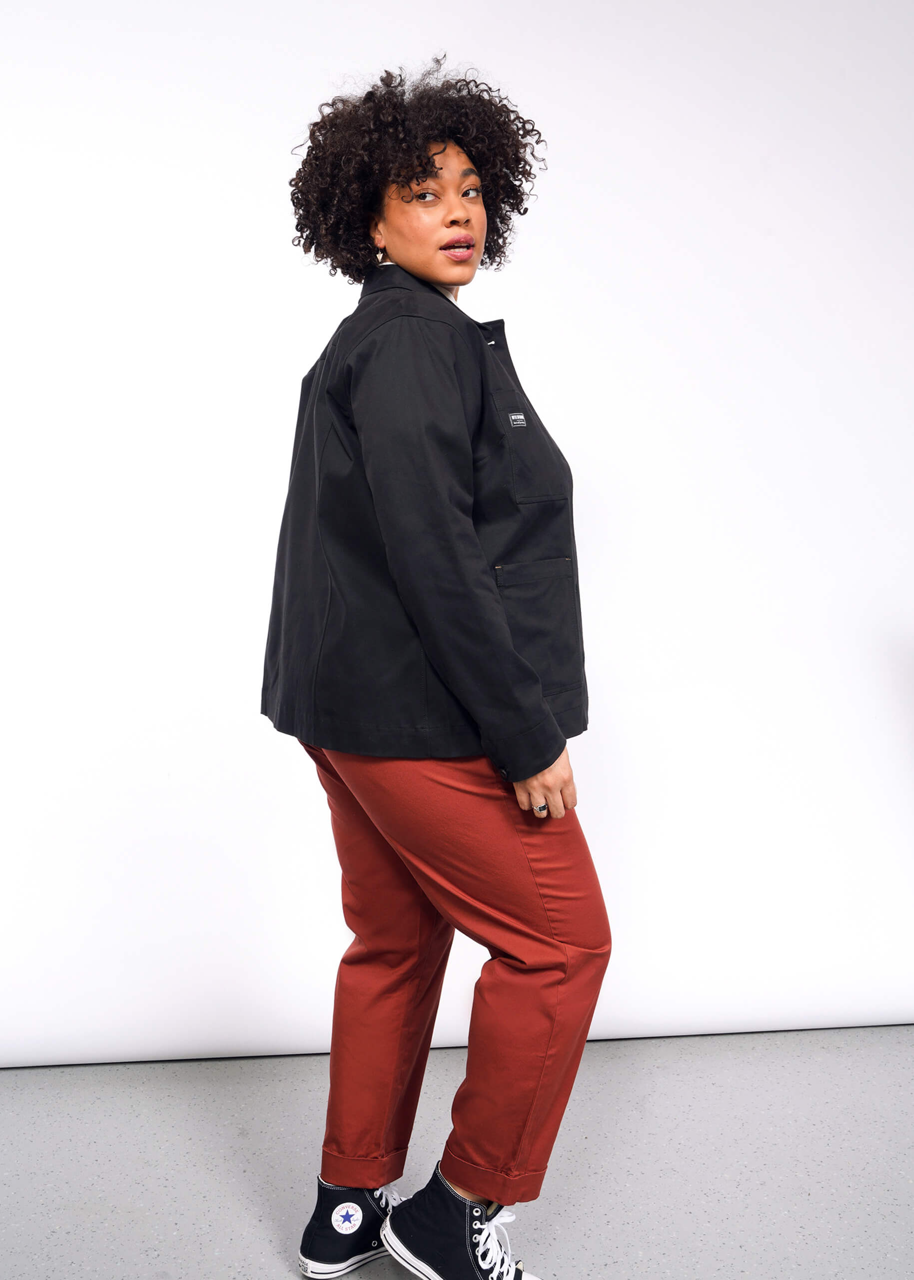 A person with curly hair stands against a white background, wearing The Essential Utility Jacket and rust-colored pants. They glance over their shoulder and wear black sneakers.