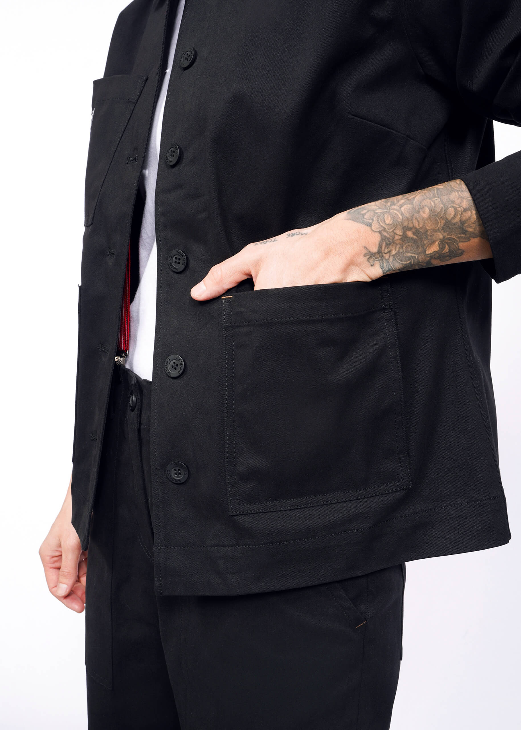 A tattooed person wears The Essential Utility Jacket, slipping their hand into its large front pocket. This black jacket over a white shirt offers stretch comfort, combining style and practicality for those valuing deep pockets and fashion details.