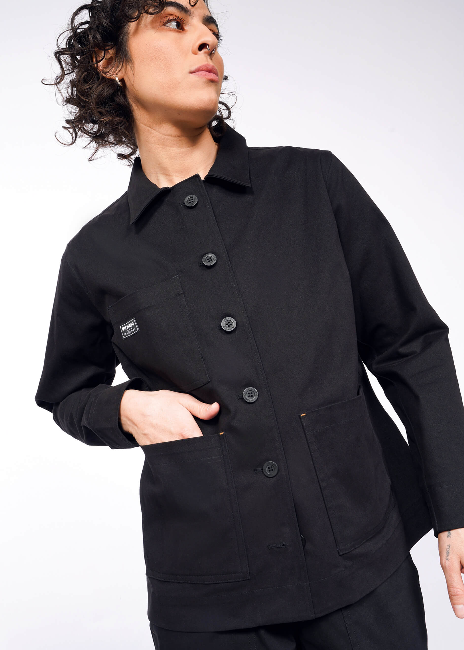 Wearing The Essential Utility Jacket in black, they stand against a plain background with their left hand in a deep front pocket. A small square label adorns the chest as their curly hair enhances the effortless blend of style and stretch comfort, while they gaze off to the side.