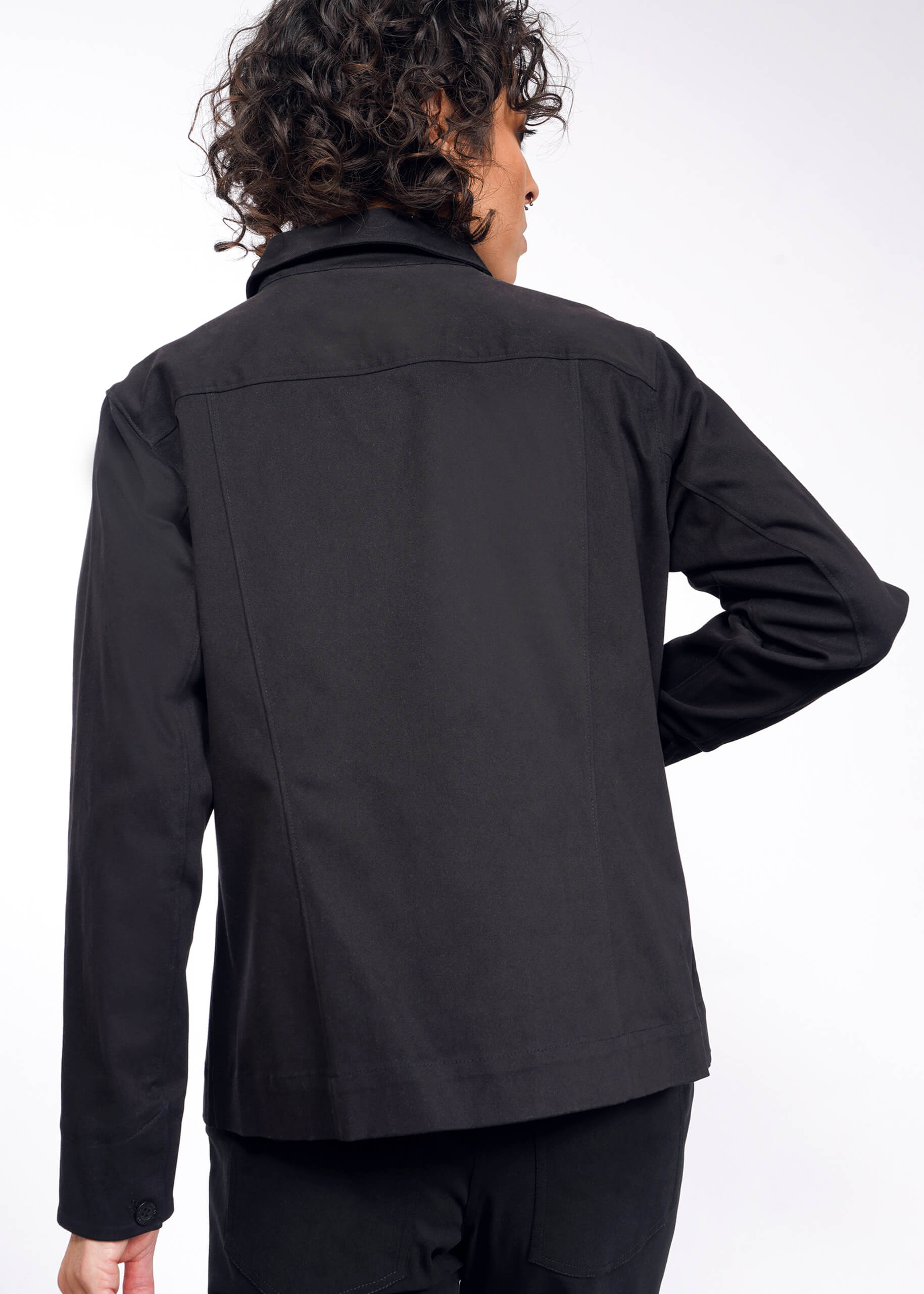 A person with curly hair, wearing The Essential Utility Jacket and facing away from the camera, stands against a plain white background. This black jacket offers stretch comfort and features deep pockets for extra functionality.