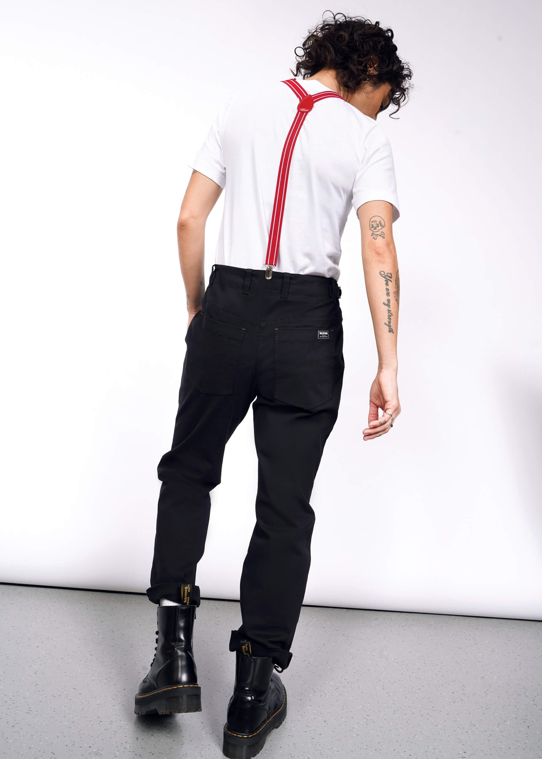 Facing away, the person wears a white t-shirt and black boots. Curly hair complements red suspenders over The Essential Utility Pant for a workwear look. Tattoos on their left arm add personal flair to the stretch cotton ensemble.