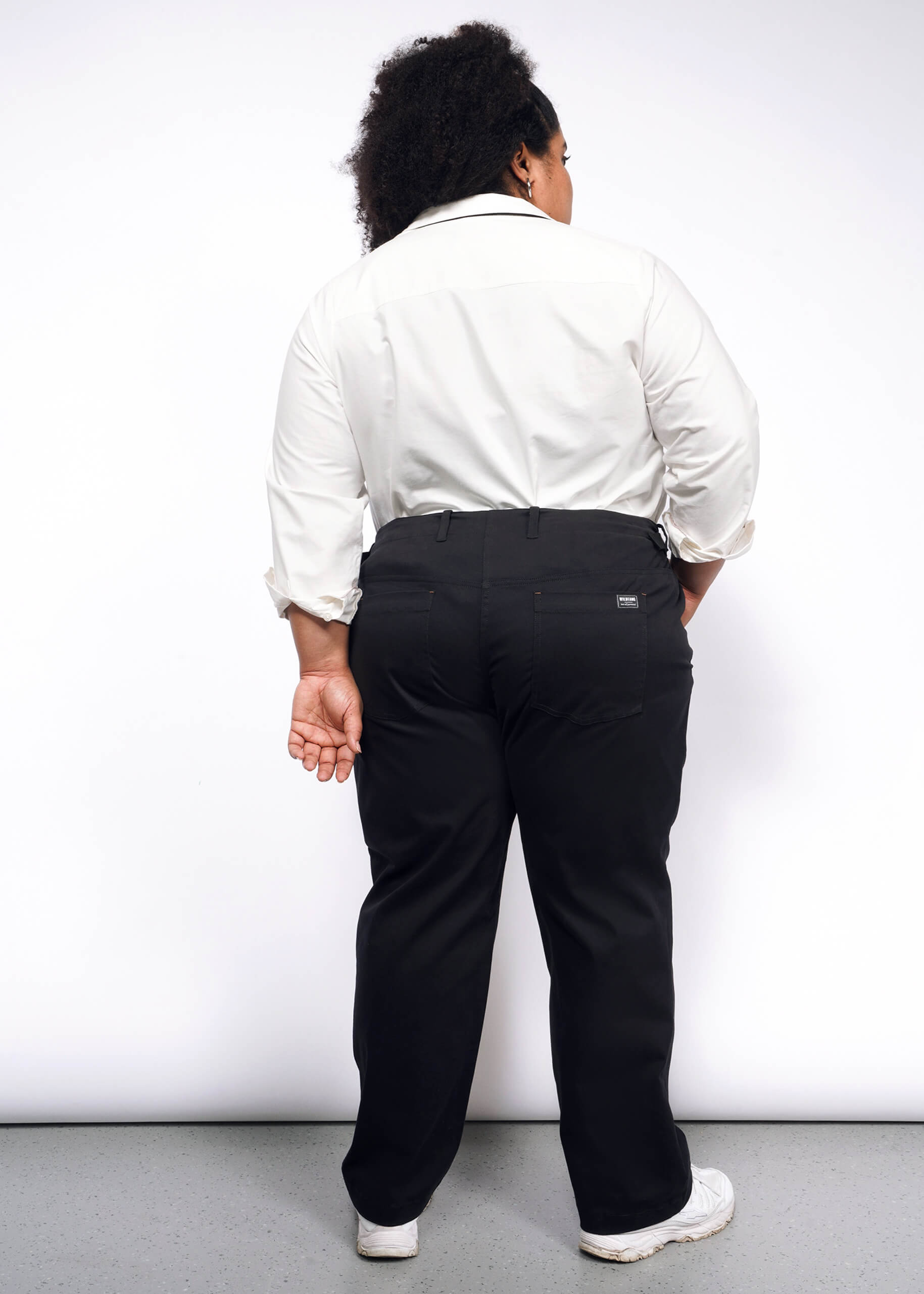 A person with long curly hair, seen from behind, wears a white shirt and black Essential Utility Pant in stretch cotton, paired with white shoes. Standing against a plain white background, their right hand is partially visible behind their back, showcasing a chic workwear style.