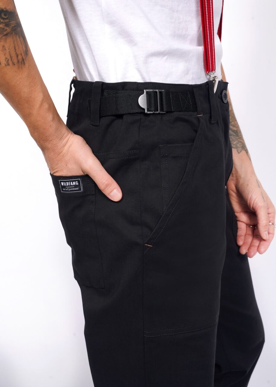 A person is shown wearing The Essential Utility Pant with a WUBIANG patch, styled in workwear fashion. Made of stretch cotton, the black pants are secured with a black belt. One hand rests in a pocket, revealing tattoos and a red strap against the plain white background.