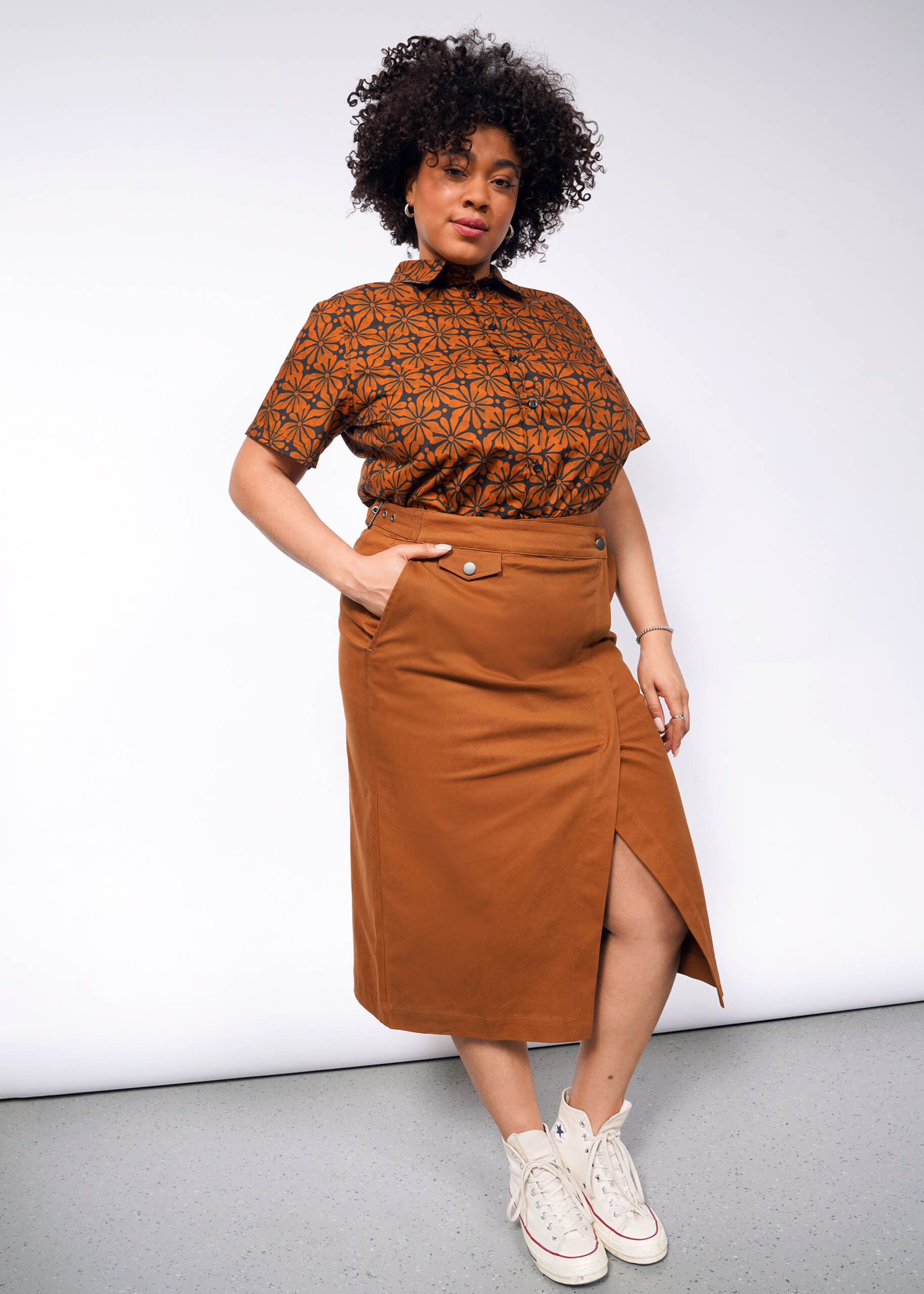 A person with curly hair poses confidently in a patterned brown shirt and The Essential Utility Wrap Skirt, featuring a zippered opening and adjustable waist buckle at the front. They are wearing white high-top sneakers, standing against a plain gray background.