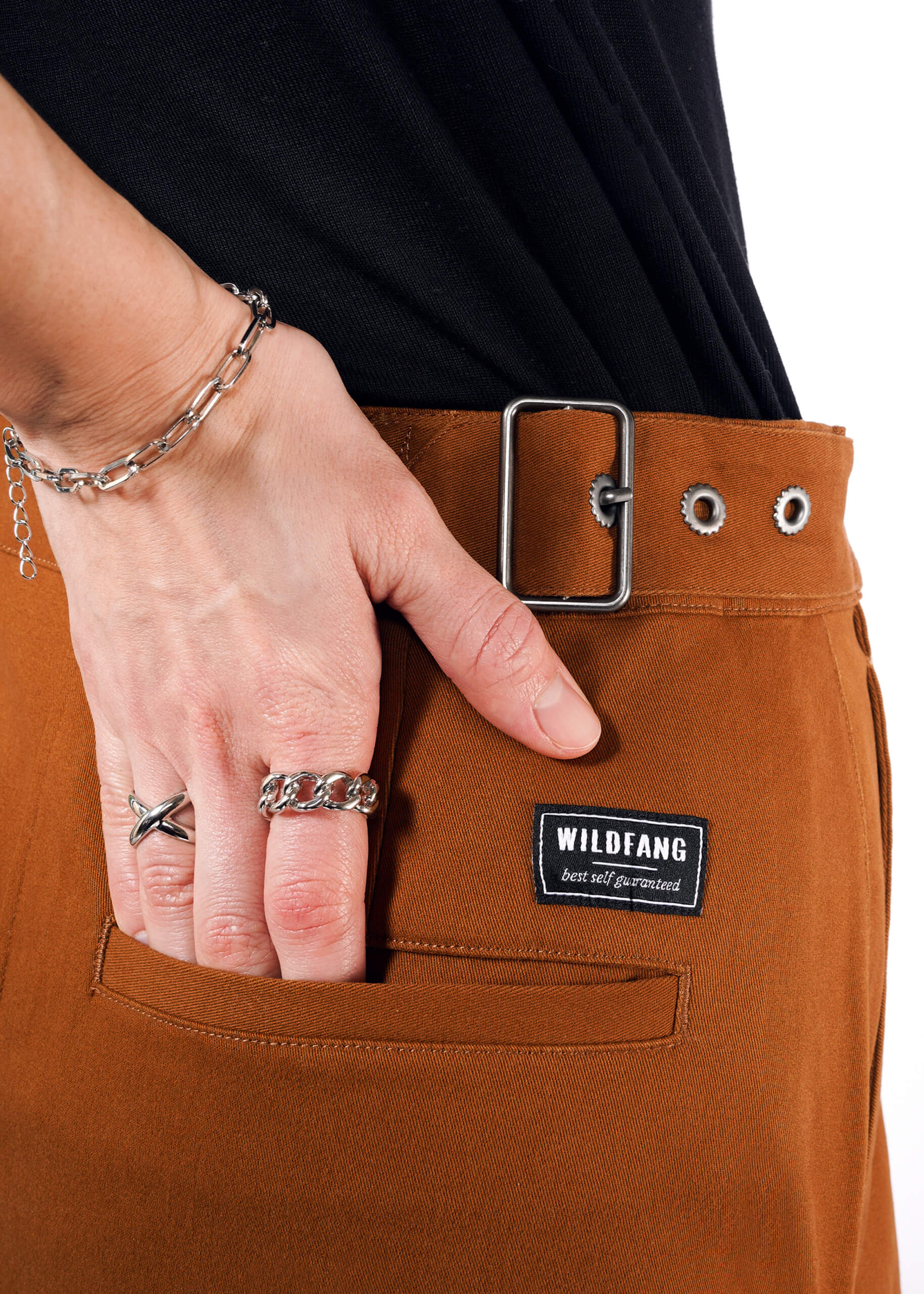 Close-up of a person in rust-colored pants featuring an adjustable waist buckle and rectangular silver clasp. A hand with silver rings and a chain bracelet rests in the back pocket, showing Wildfang branding. The outfit is completed with a sleek black shirt. Product: The Essential Utility Wrap Skirt.
.