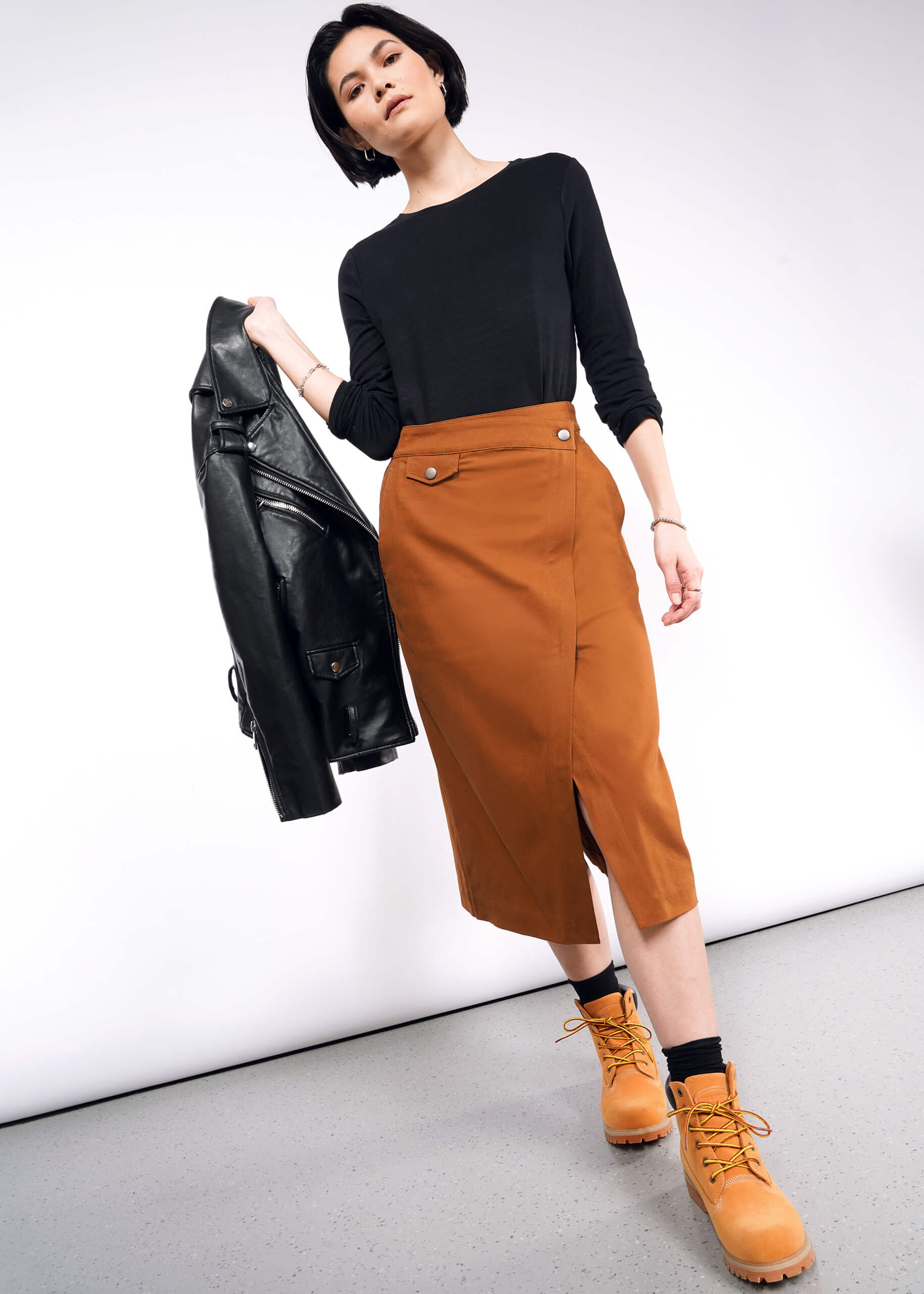 A person poses confidently in a black long-sleeve top and The Essential Utility Wrap Skirt, an orange midi skirt with an adjustable waist buckle, paired with matching orange boots. They hold a black leather jacket with a zippered front against a plain white background.