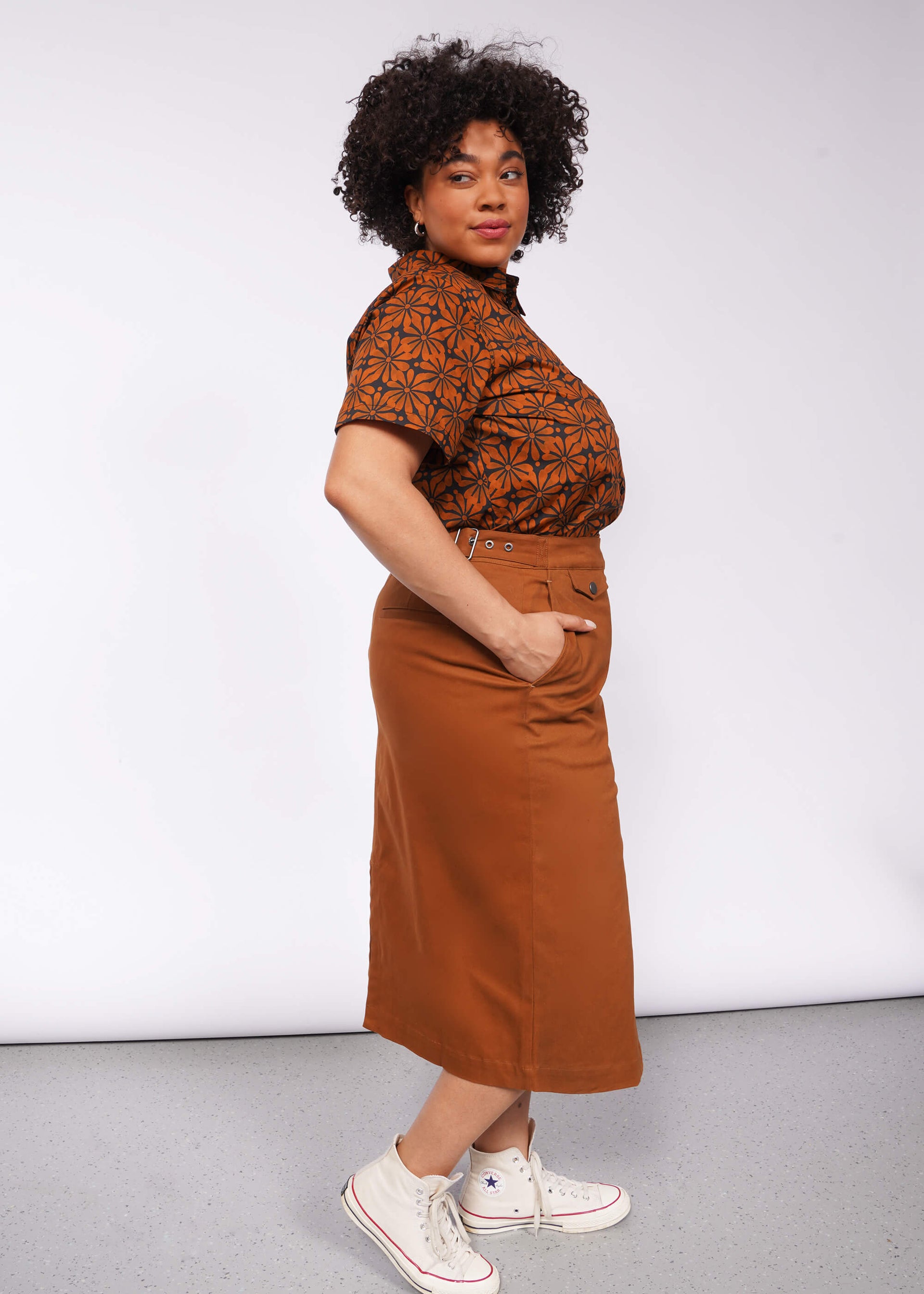 A person with curly hair stands confidently in a patterned short-sleeve shirt and The Essential Utility Wrap Skirt, featuring an adjustable waist buckle, paired with white sneakers. The background is plain gray.