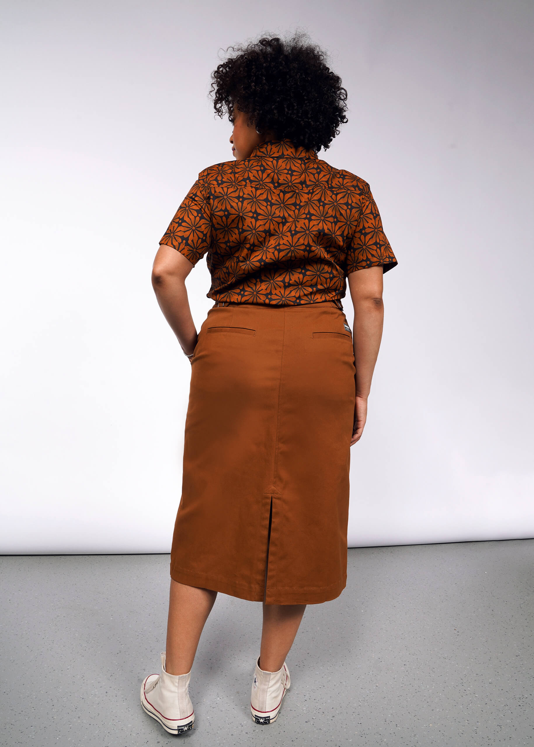 A person with curly hair stands with their back to the camera, wearing a brown geometric-patterned shirt and The Essential Utility Wrap Skirt, which has an adjustable waist buckle. They are in white sneakers on a gray floor against a white backdrop.