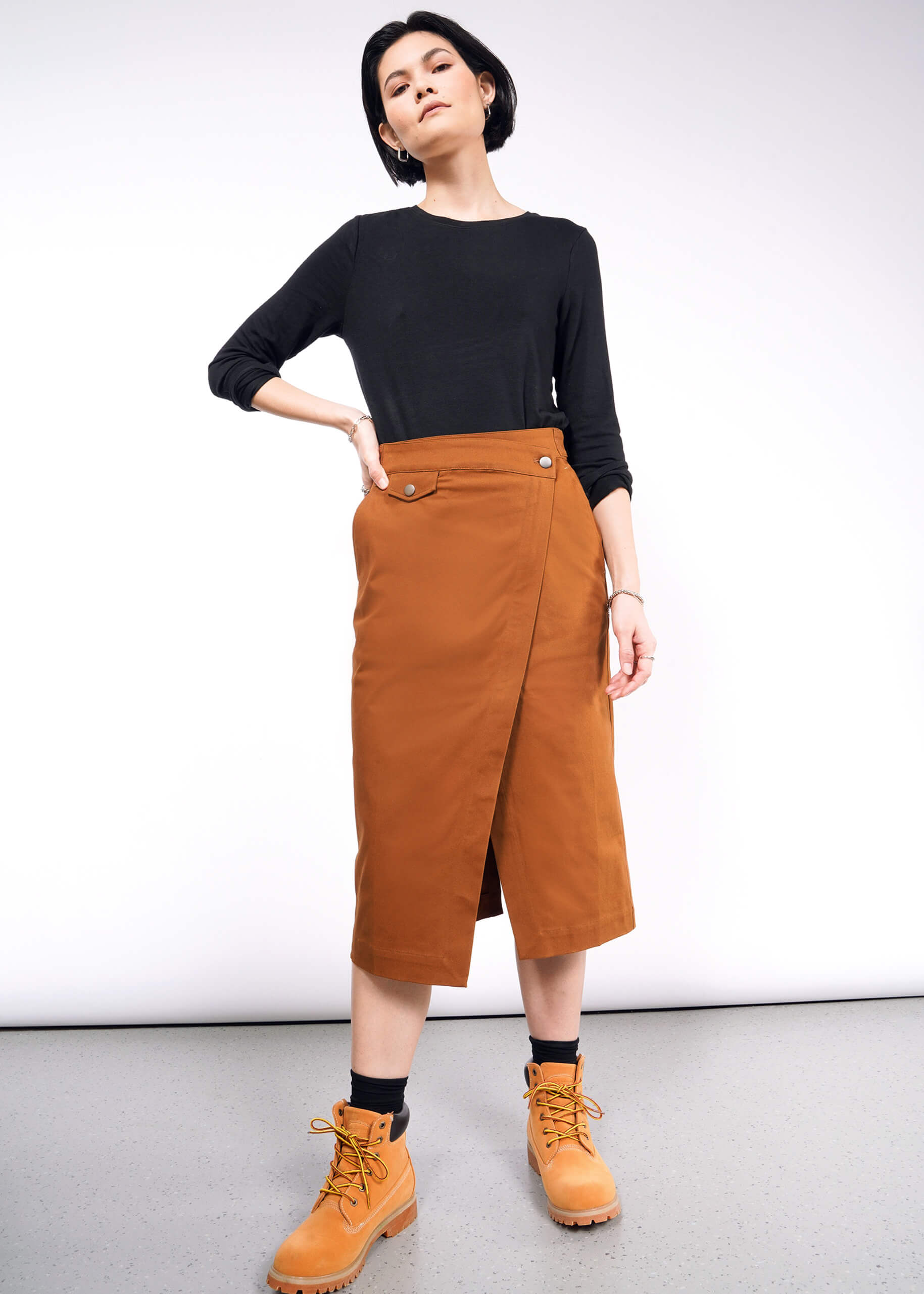 A person dressed in a black long-sleeve top, The Essential Utility Wrap Skirt with button details and an adjustable waist buckle, complemented by tan ankle boots. Their short hair is styled as they confidently pose with one hand on their hip in a minimalist space.