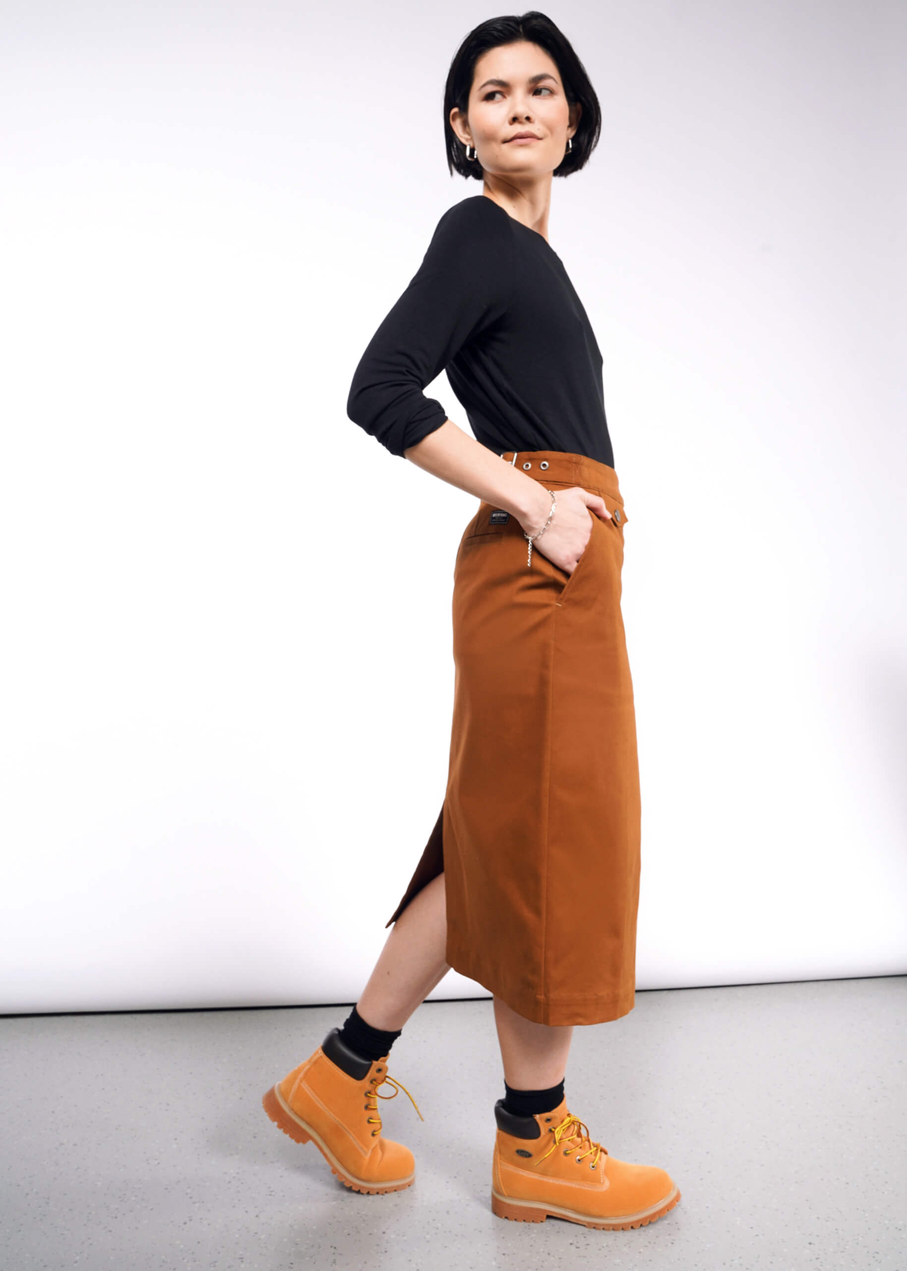 A person with dark hair is wearing a black shirt, The Essential Utility Wrap Skirt in brown with a zippered opening, and mustard yellow boots. They stand in profile against a plain white background, hands in pockets, maintaining a neutral expression.