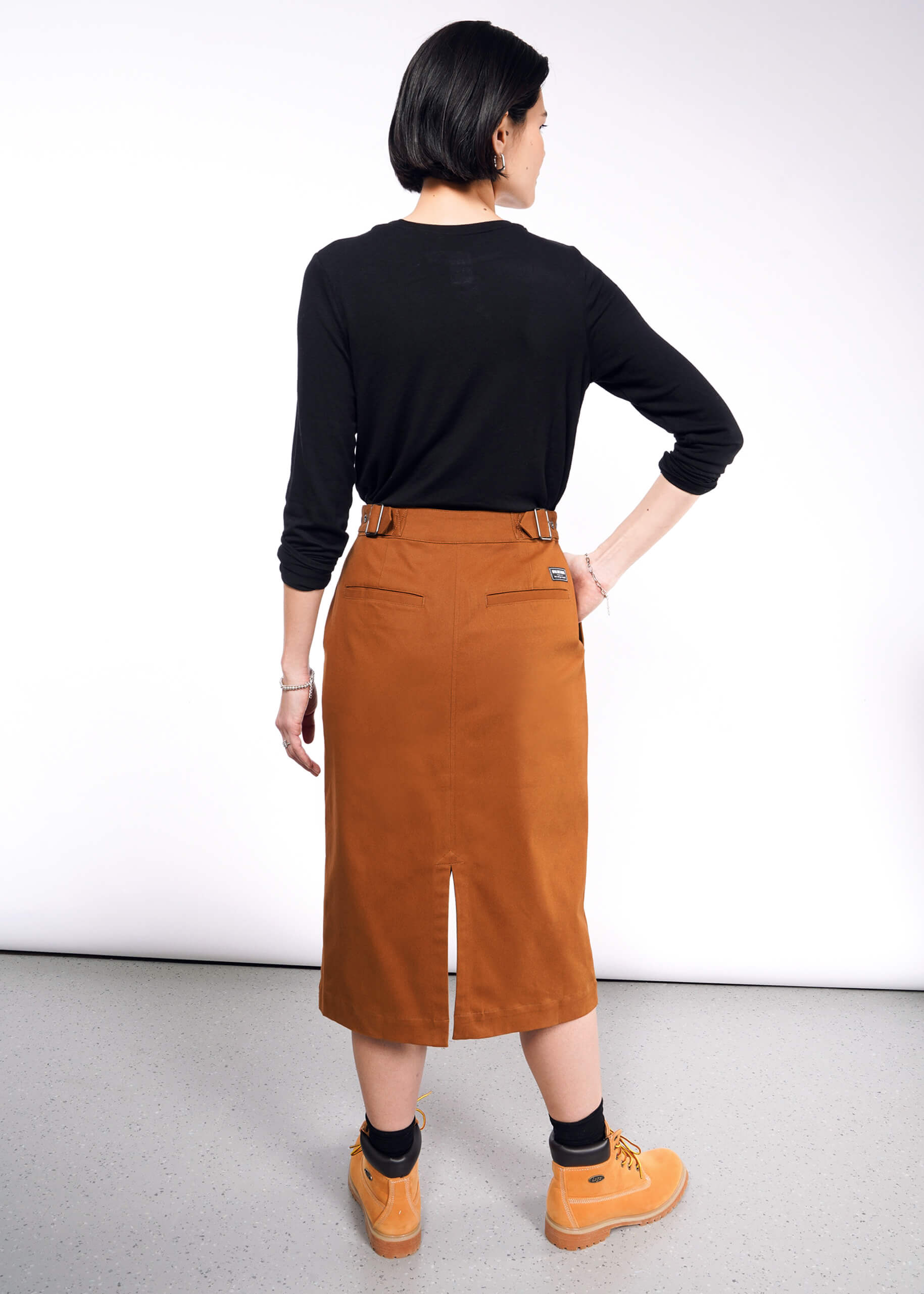 A person with short black hair wears a black long-sleeve top and The Essential Utility Wrap Skirt, a brown midi with back slit and adjustable waist buckle. Facing away from the camera, they sport tan boots on a light gray floor against a white wall.