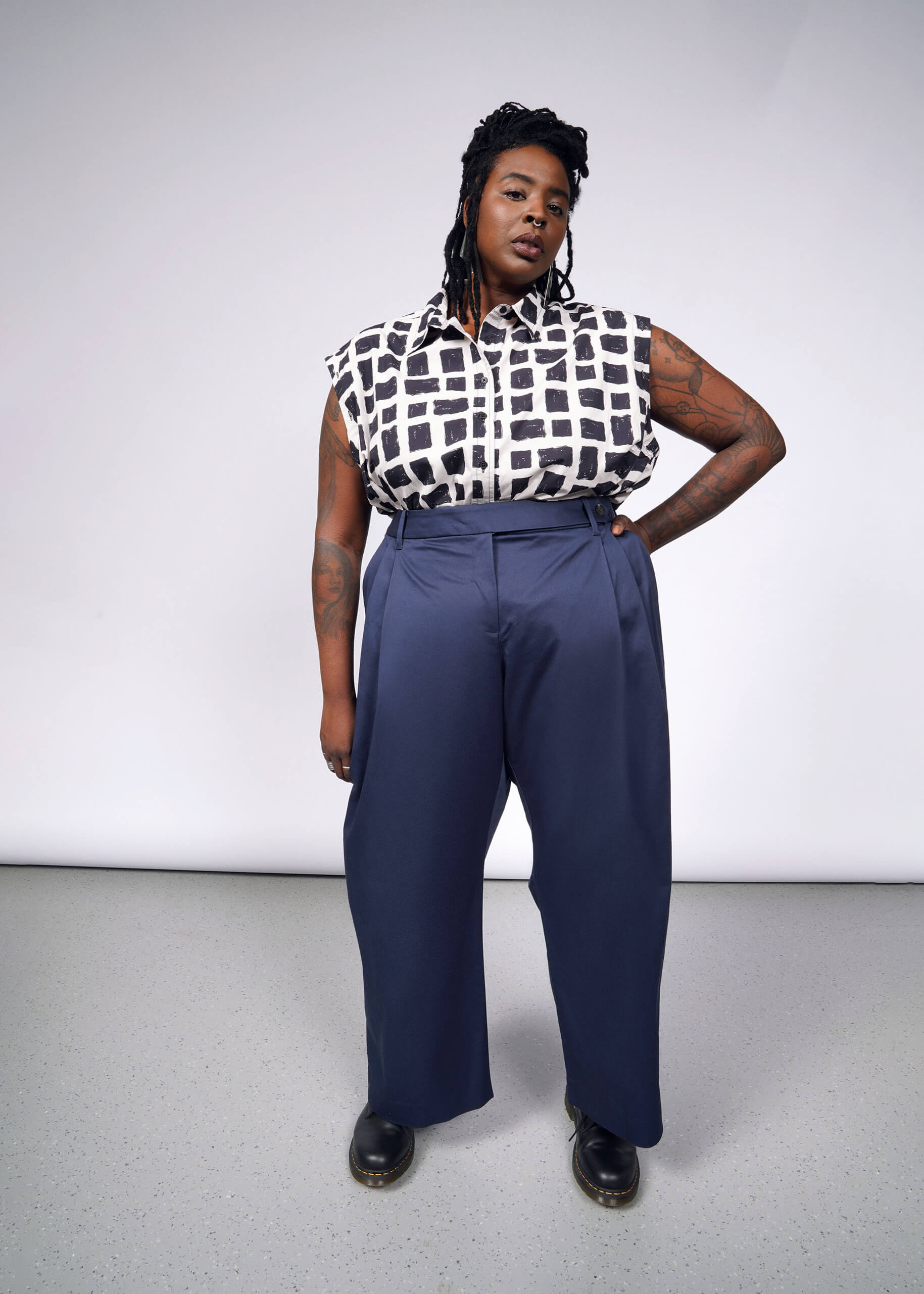 Standing confidently on a light gray floor, this individual wears The Essential Wide Leg Trouser, a high-waisted, wide-leg navy blue piece made from cotton stretch fabric, paired with a sleeveless black-and-white square patterned top. Long braided hair and sleek black shoes complete the look.