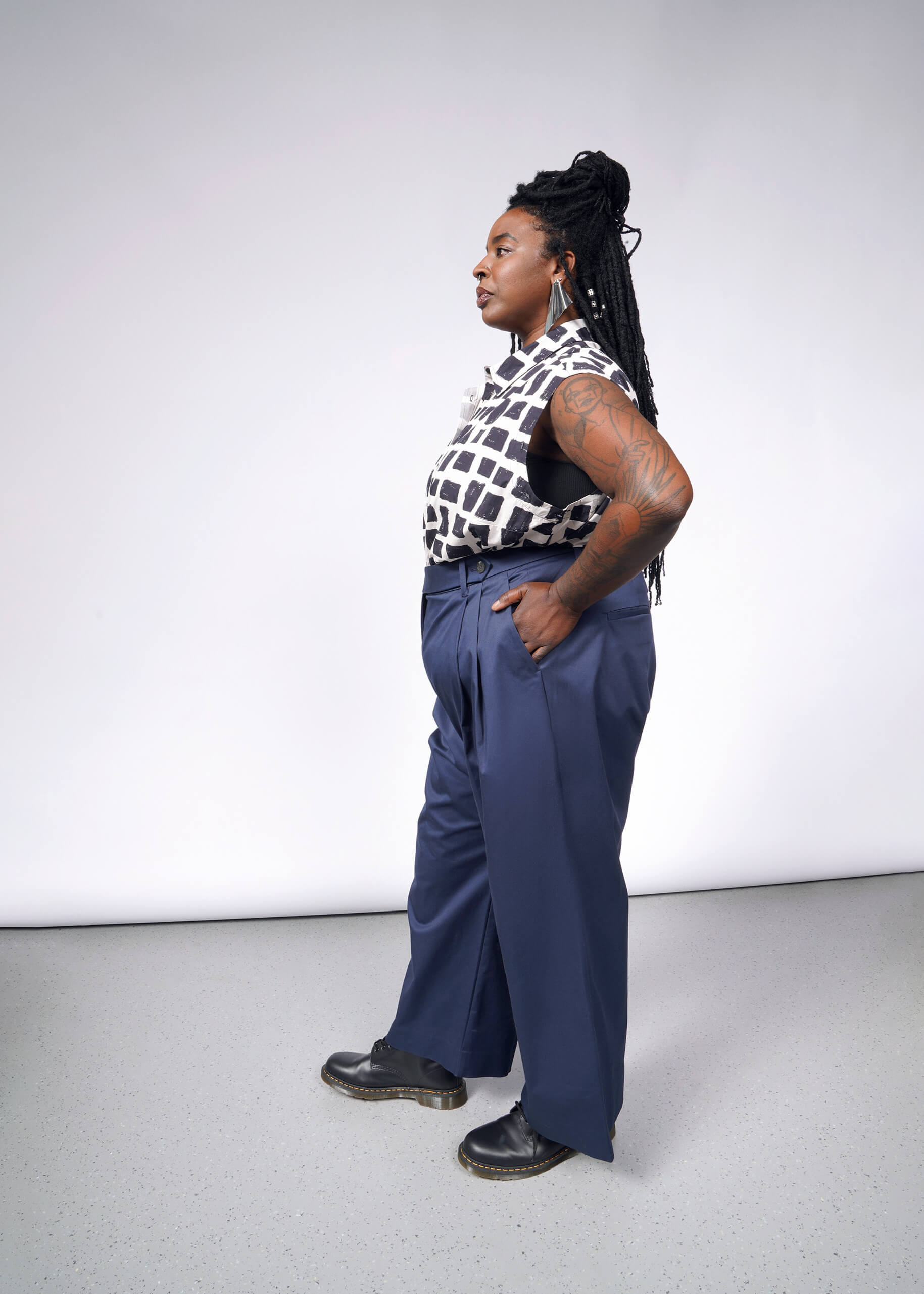 A person with long braided hair stands sideways against a gray background, wearing The Essential Wide Leg Trouser in blue cotton stretch fabric, paired with a sleeveless black and white patterned top. Black shoes complete the look, accentuated by tattoos on their arms.