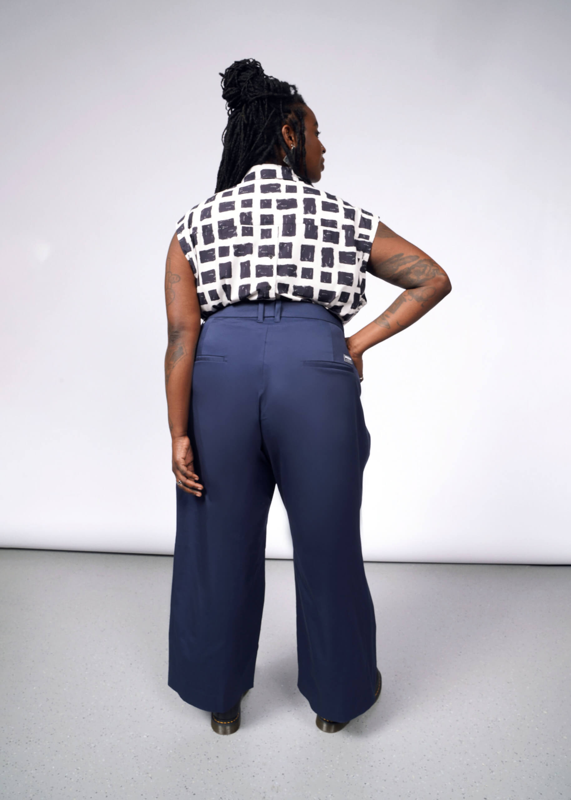 Facing away from the camera against a plain gray backdrop, a person dons The Essential Wide Leg Trouser in navy blue cotton stretch, paired with a sleeveless white and black checkered top. Their right hand rests on their hip.