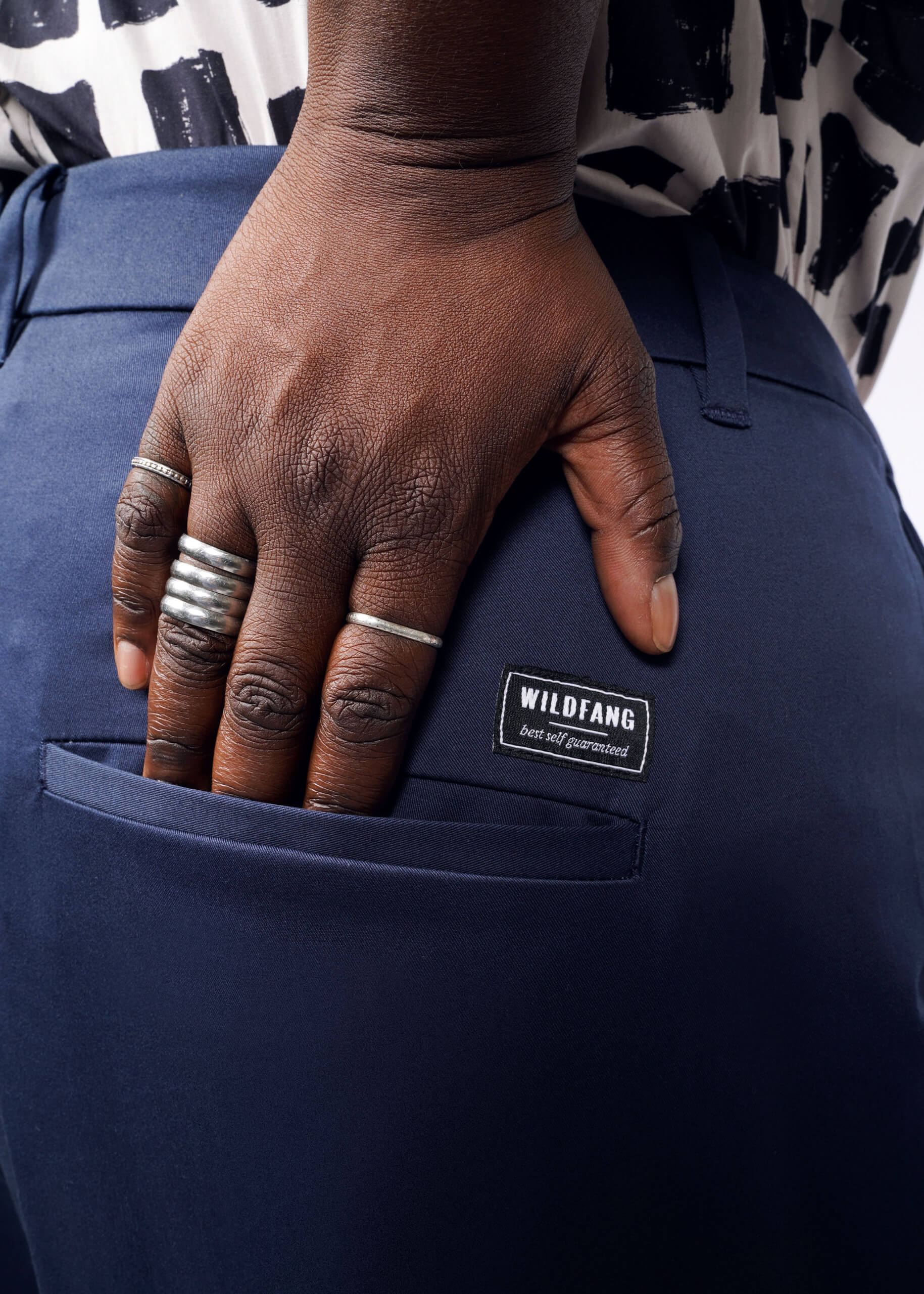 A hand rests on the back pocket of Wildfangs The Essential Wide Leg Trouser, high-waisted and made from dark blue cotton stretch fabric. The person sports multiple silver rings and a black and white patterned top.