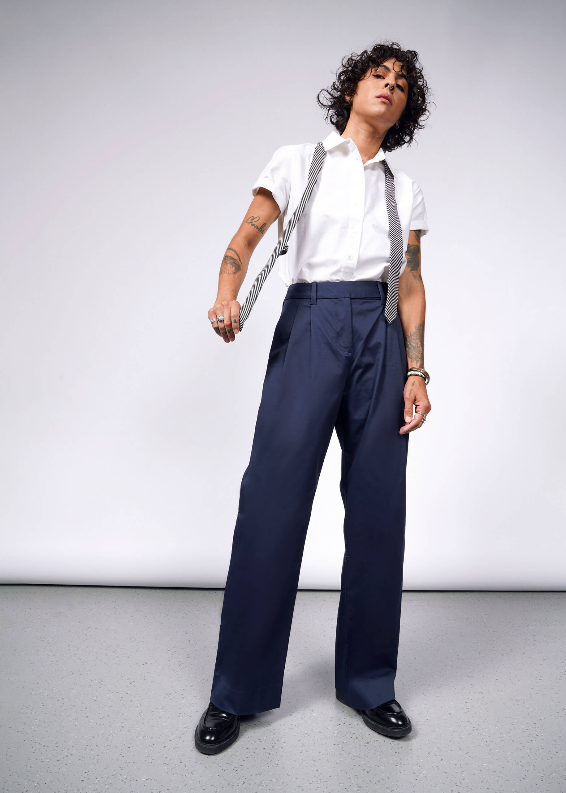 A person with curly hair confidently stands against a plain background, showcasing tattoos on their arms. Theyre wearing a white short-sleeve shirt and navy The Essential Wide Leg Trouser, made from cotton stretch fabric, holding suspenders, and paired with black shoes.