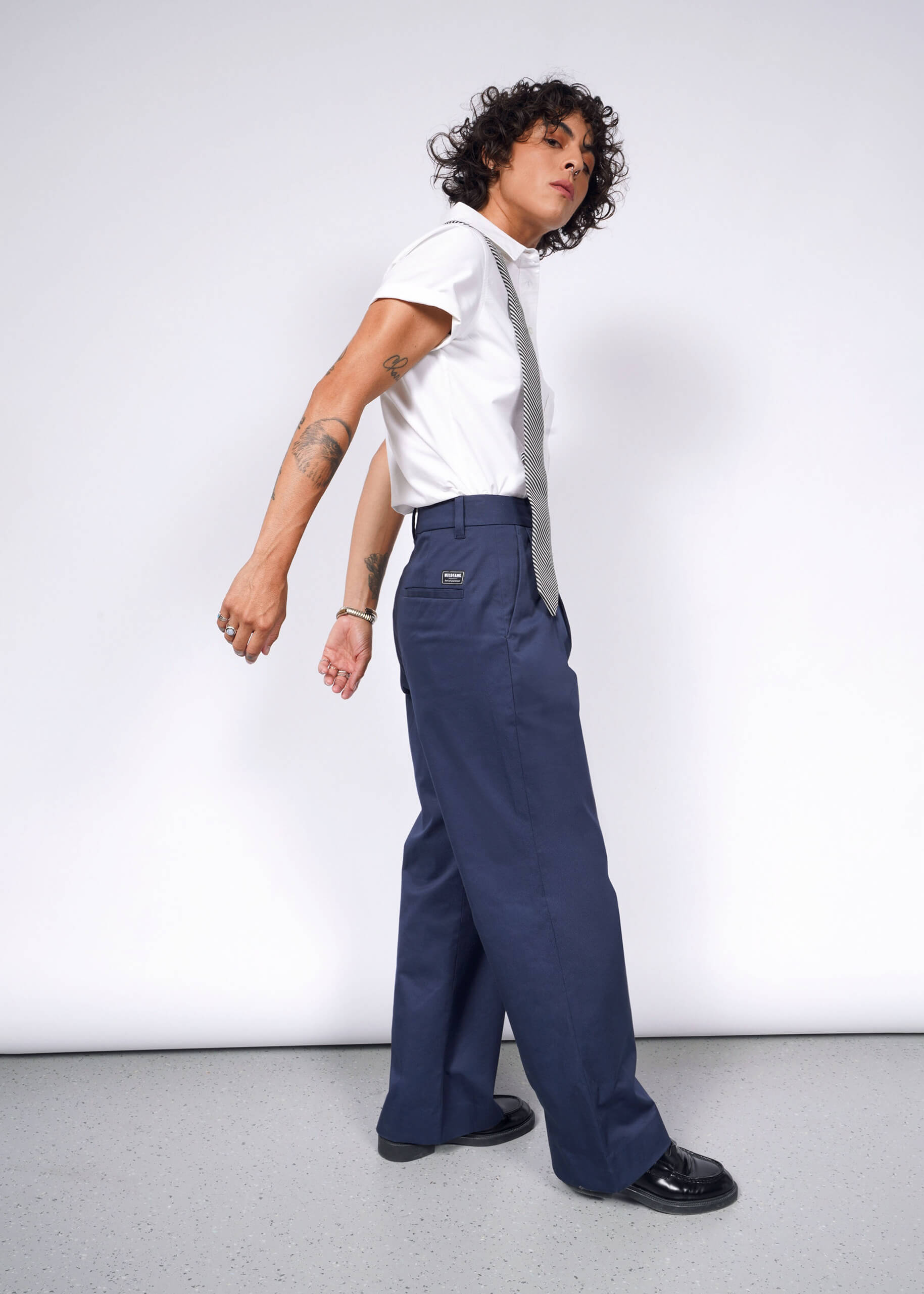 A person with curly hair, a white t-shirt, and a silver tie confidently poses in The Essential Wide Leg Trouser made from cotton stretch fabric. Tattoos adorn their arms as they stand in black shoes against a plain white background.