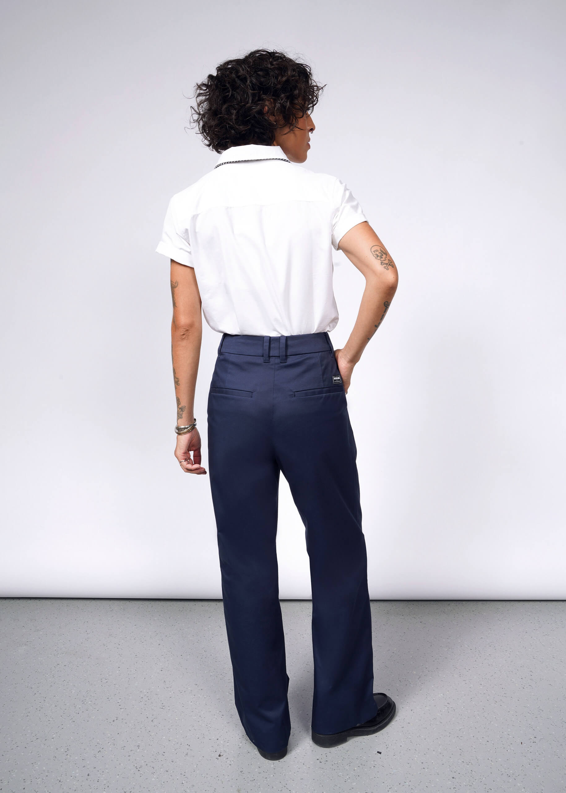 A person with short, curly hair stands facing away in a white short-sleeve shirt and The Essential Wide Leg Trouser in navy blue cotton stretch. Tattoos adorn their arms as their right hand rests on their hip against a plain white background.