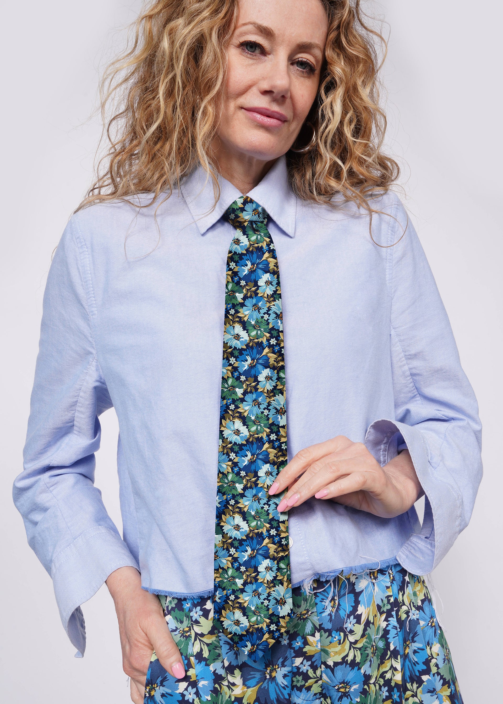 Person with blonde curly hair looks at camera wearing blue button up shirt, floral blue and green patterned tie, and matching pants.