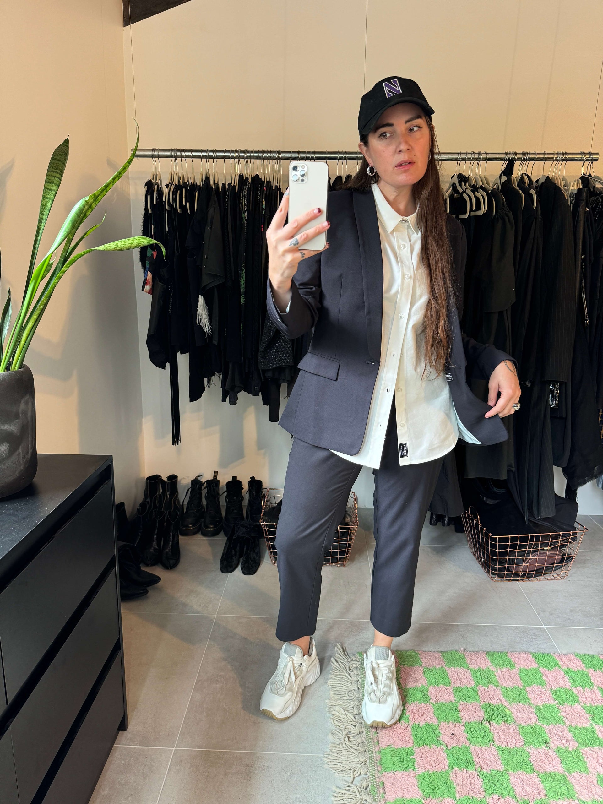 Wearing The Empower Slim Crop Pant, a dark suit, and a white shirt, the person captures a mirror selfie. Sporting a black cap and sneakers amidst black clothing racks, they stand on a green and pink checkered rug with a plant to the left.