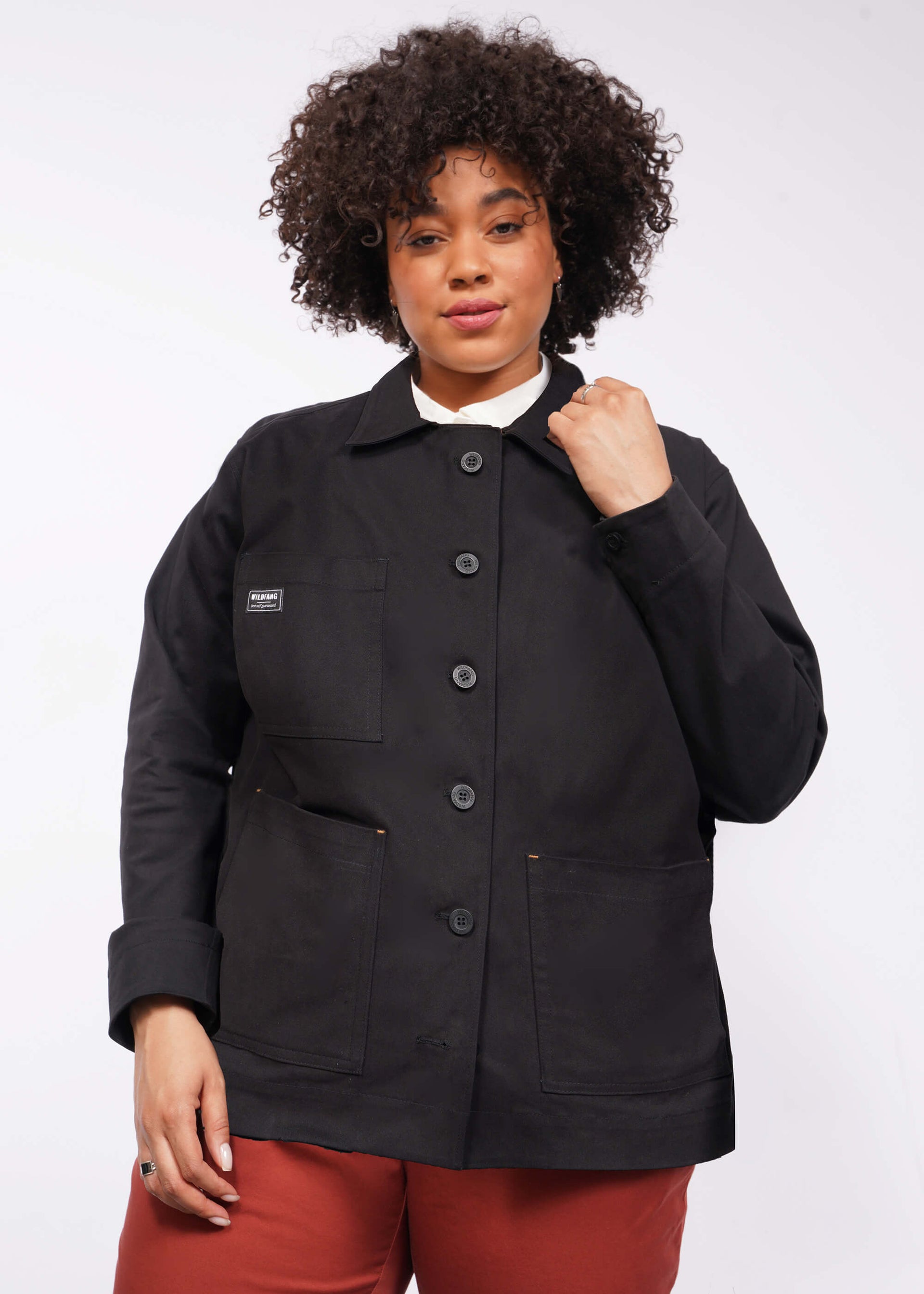 The person is wearing The Essential Utility Jacket, a black utility jacket with deep front pockets, paired with red pants. Theyre posing against a white background, have curly hair, and are looking at the camera with a slight smile.