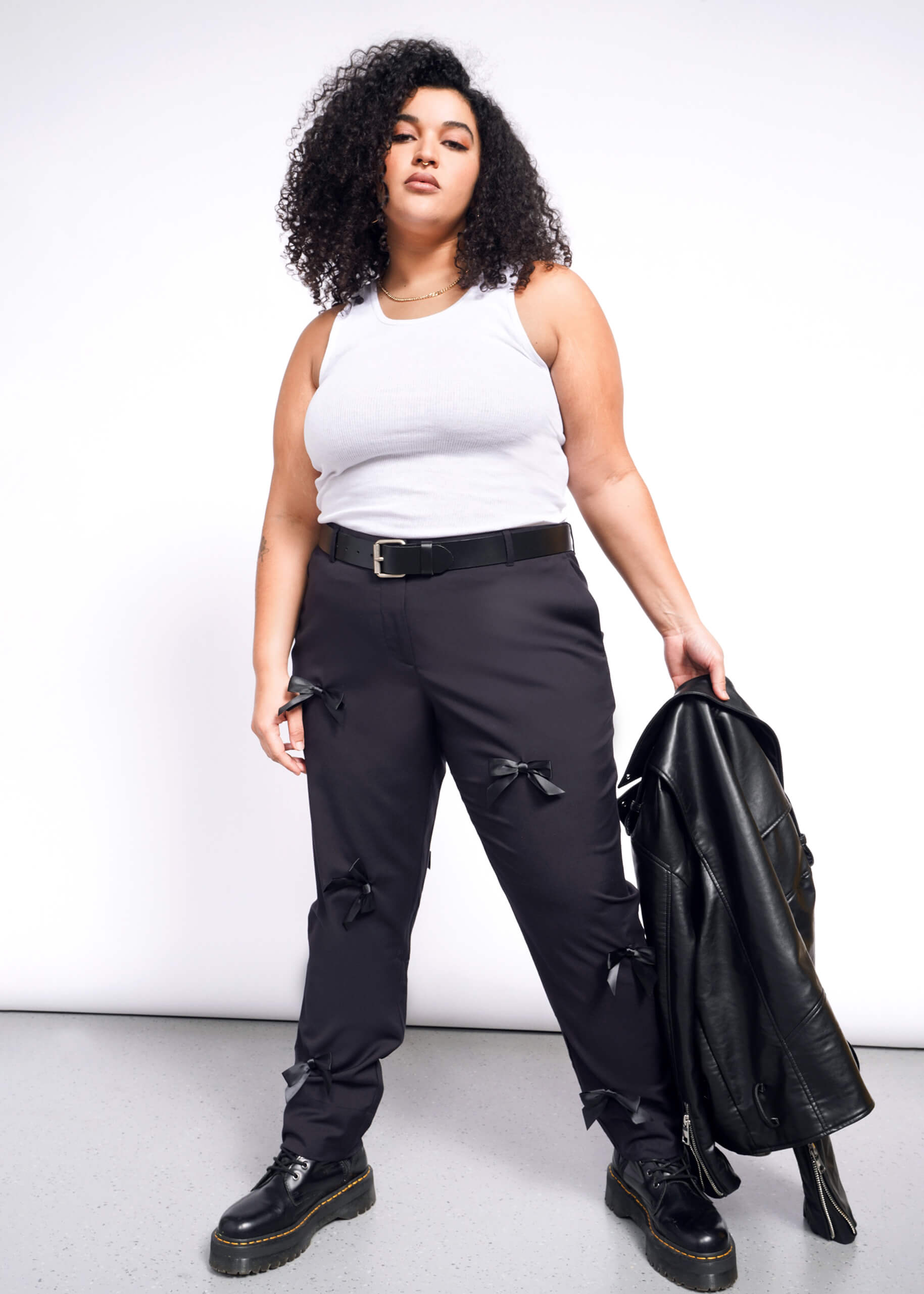 A curly-haired woman in a white tank top and The Empower Bows Trouser stands confidently, holding a leather jacket. Her ultra-lux style is completed with black boots against a plain background.