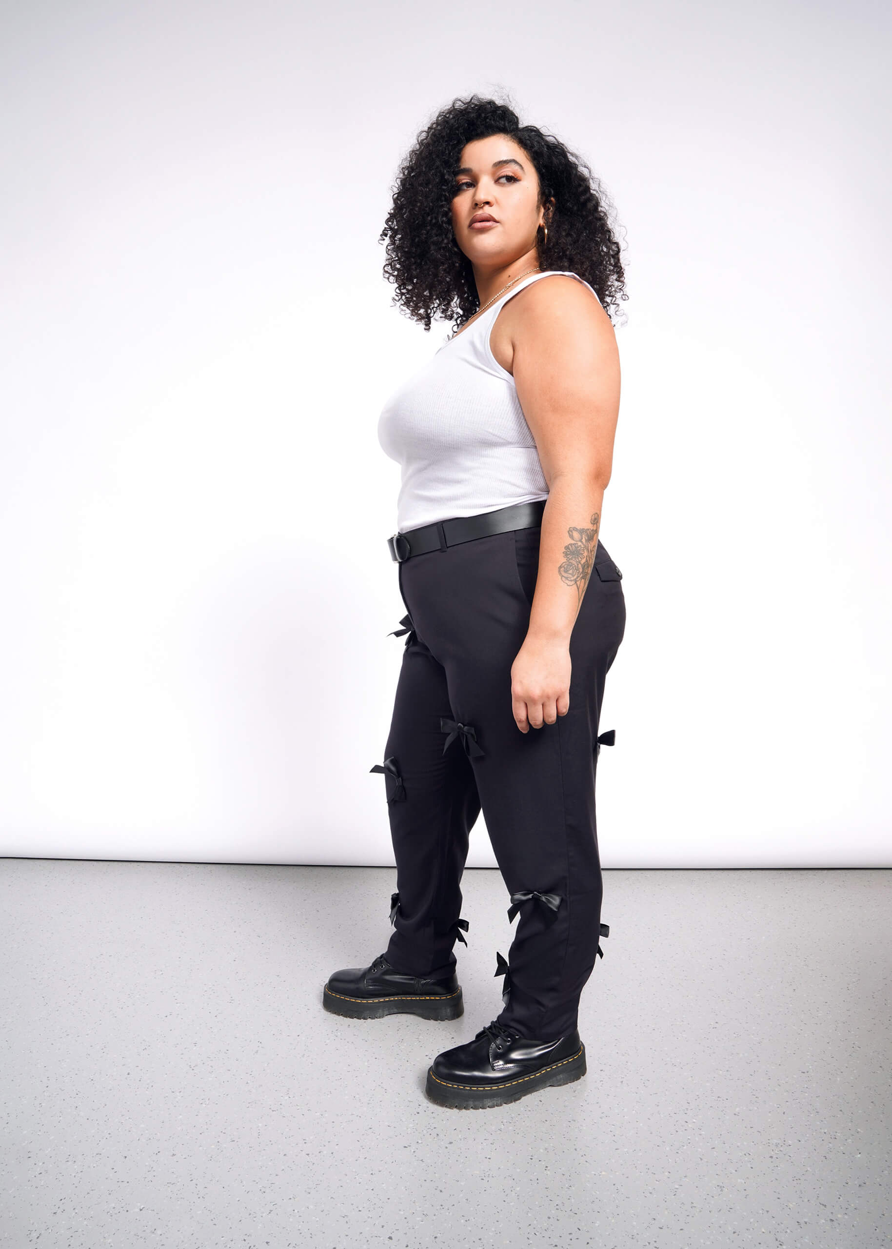 A person with curly hair in a white tank top and The Empower Bows Trouser stands on a speckled floor against a white background. Their right arm features a tattoo, and black shoes complete the chic ensemble.