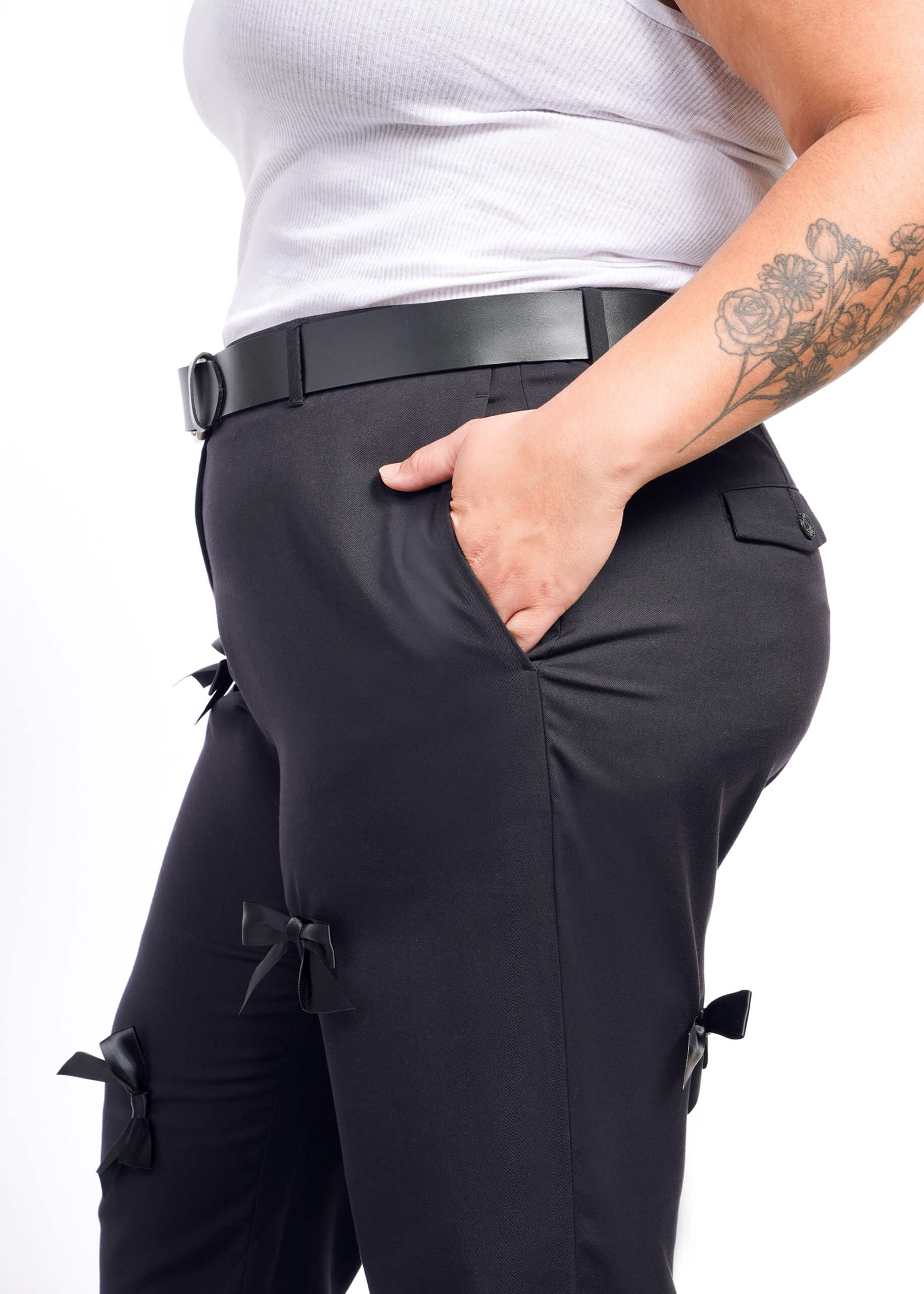 A person wearing The Empower Bows Trouser with a black belt and white tank top is seen against a crisp white background. One hand in their pocket reveals a floral tattoo, exuding an ultra-lux feel.