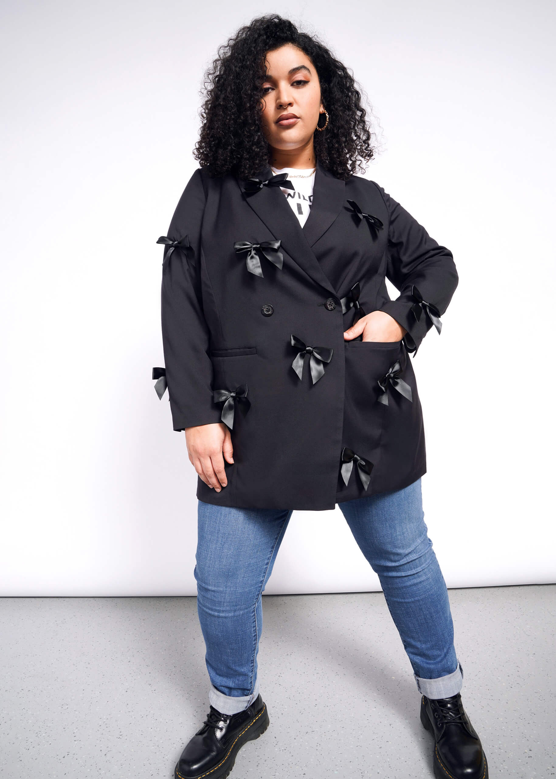 A person with curly hair confidently stands against a white background, wearing The Empower Bows Double Breasted Blazer over a white shirt, paired with blue jeans and black shoes.