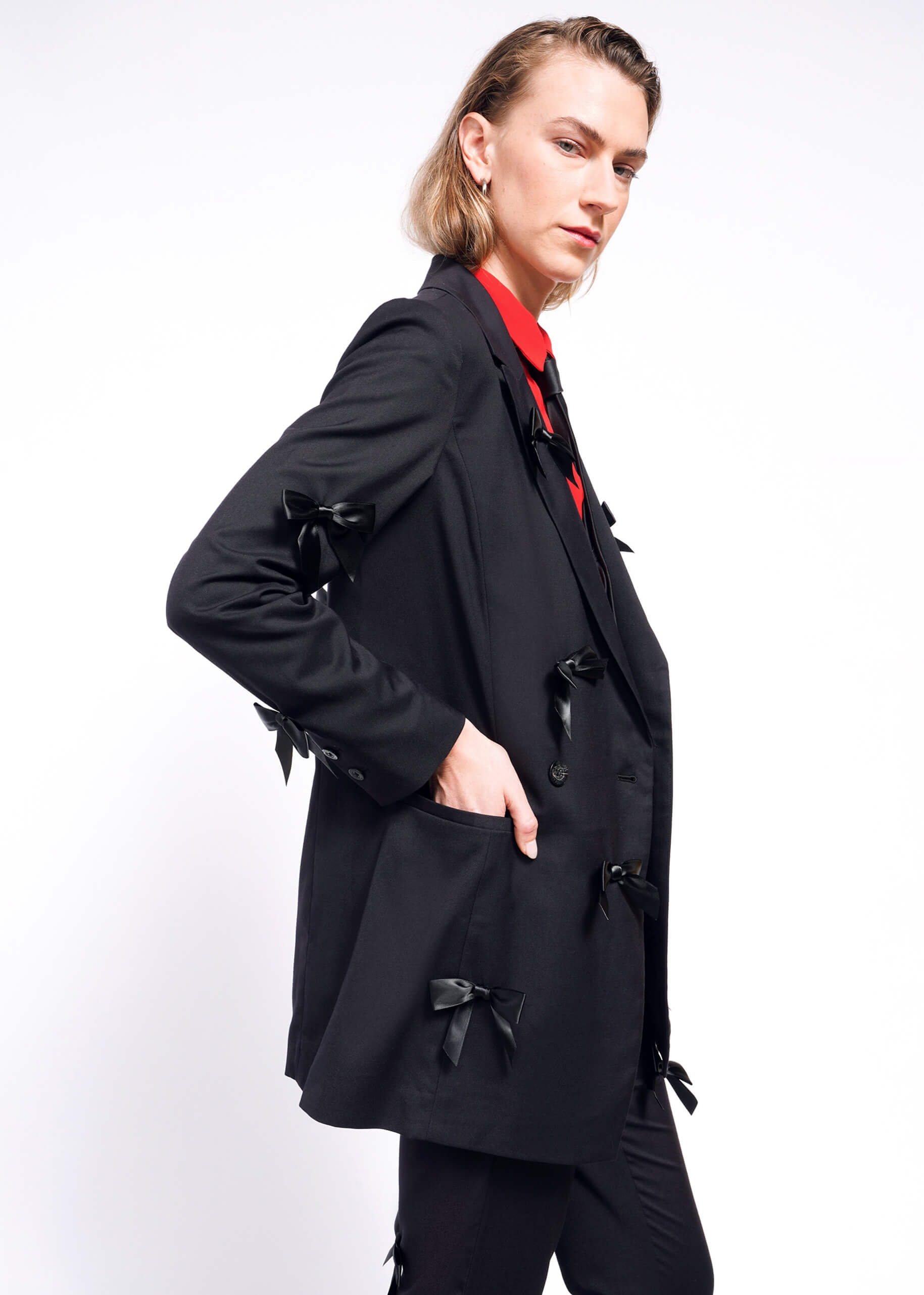 Wearing The Empower Bows Double Breasted Blazer, adorned with black bows and a red shirt, a person with shoulder-length hair stands sideways against a white background. With their hand in the blazer pocket, they exude confidence.