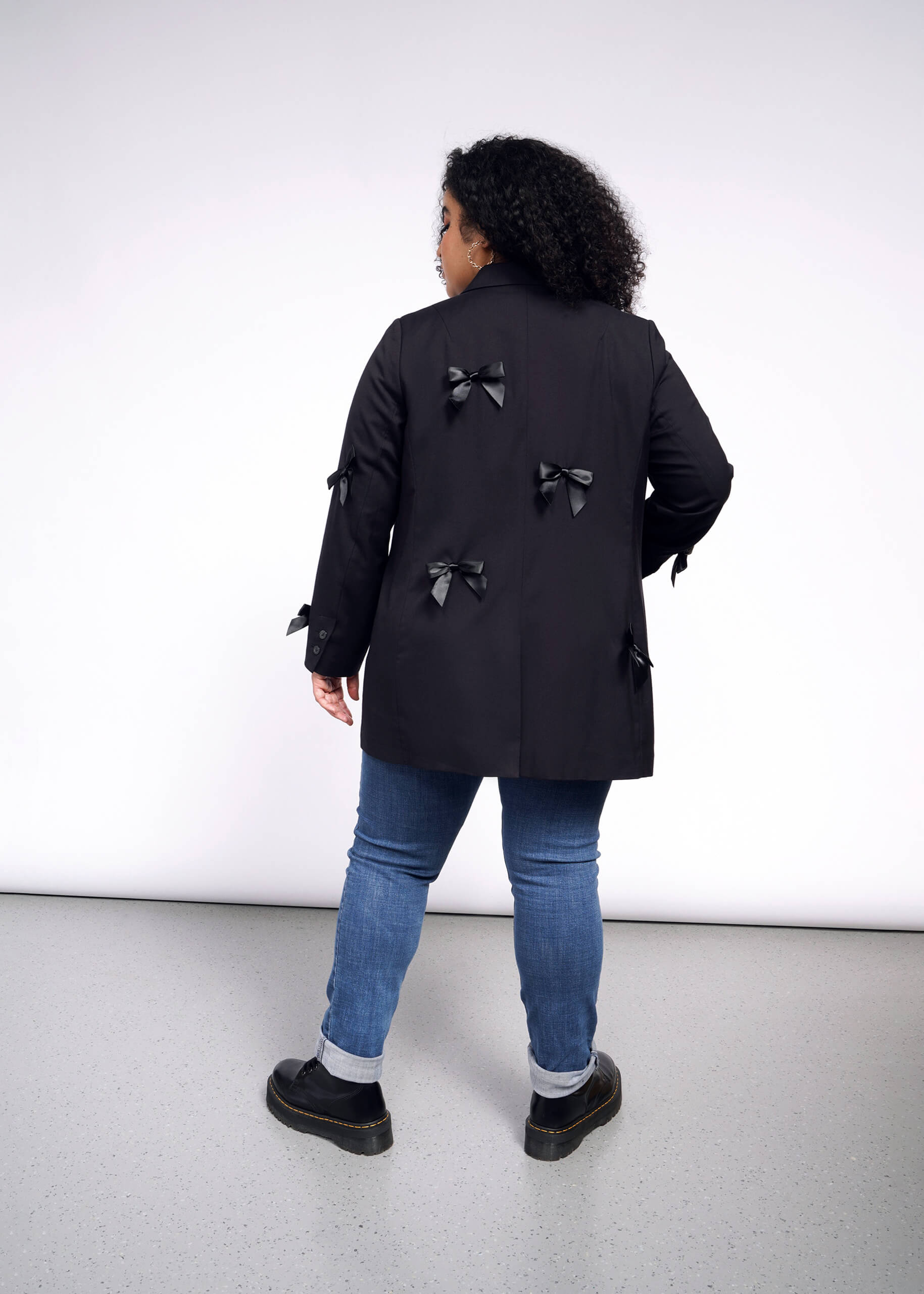 A person with curly hair faces away, wearing an oversized black coat adorned with multiple bows on the back and sleeves. They pair it with blue jeans and black shoes, standing on a light gray floor against a white background. The ensemble flows elegantly like The Empower Bows Double Breasted Blazer.