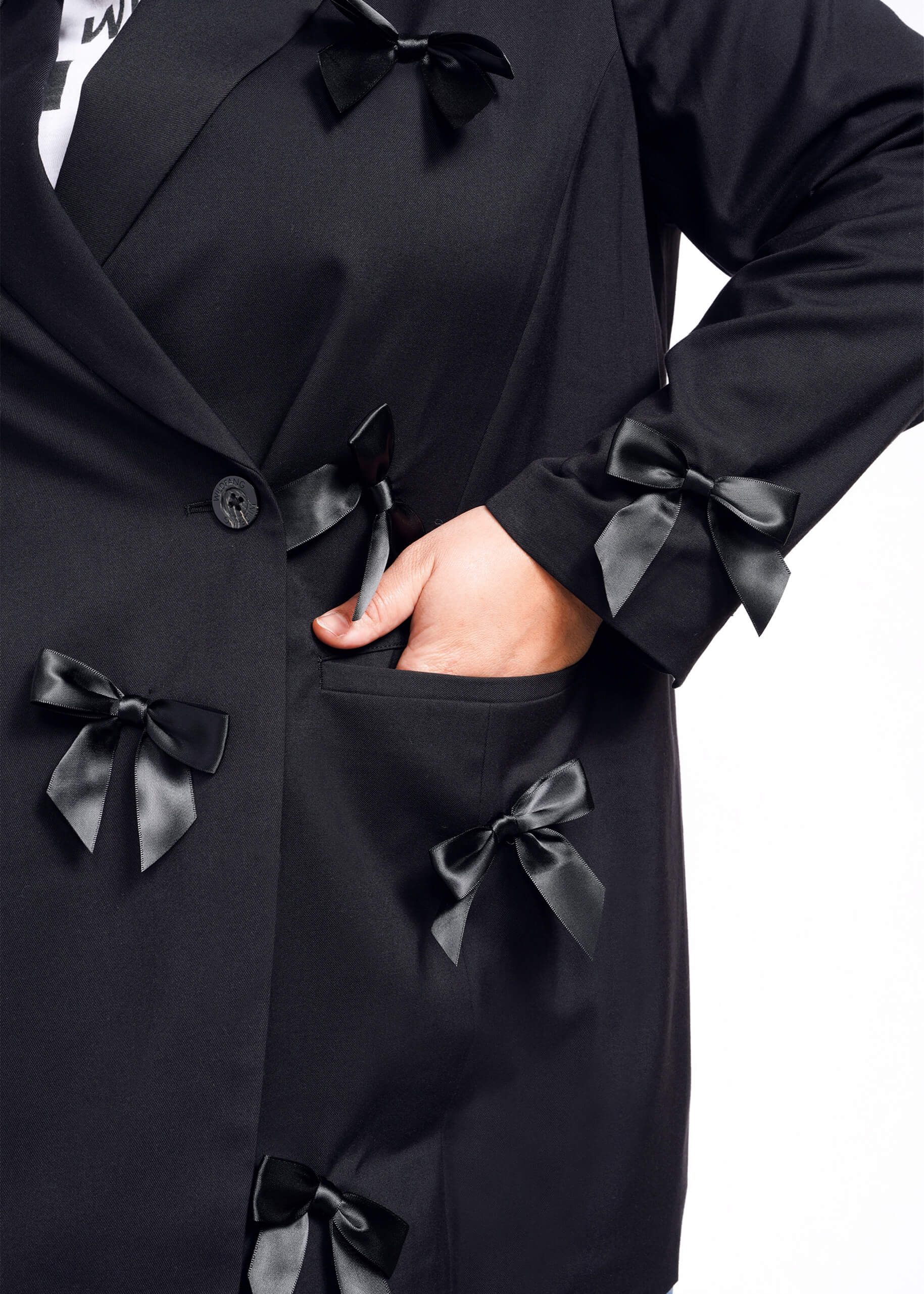 A person wearing The Empower Bows Double Breasted Blazer is photographed against a white background. The tailored oversized coat features black satin bows and drapes elegantly, with their left hand resting in one of its pockets.