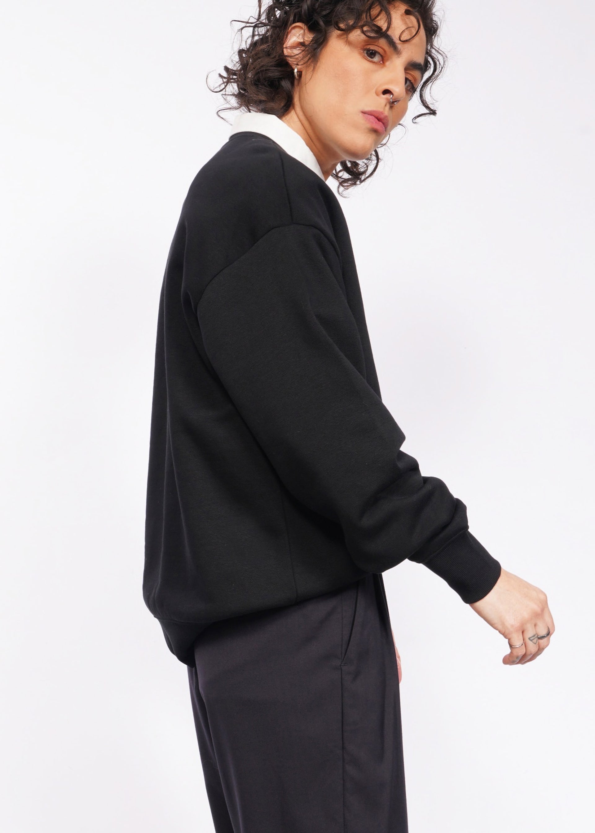 Someone with curly hair, sporting the Wild Feminist Fleece Crew in black and dark pants, poses against a light backdrop. Their expression is neutral, gazing slightly to the side.