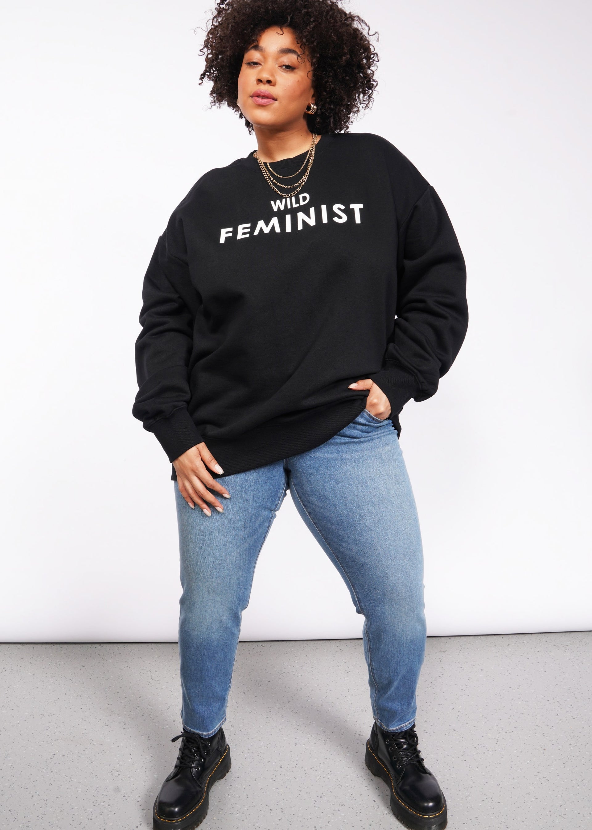 A curly-haired individual confidently stands against a white backdrop wearing the Wild Feminist Fleece Crew in black, paired with light blue jeans and black boots.