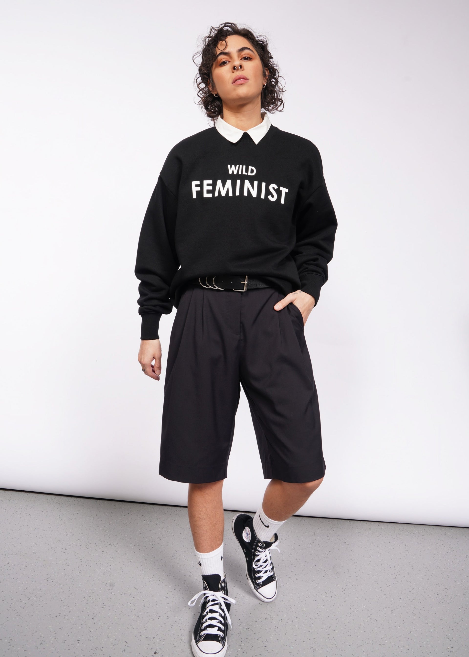 A person with short curly hair is wearing the Wild Feminist Fleece Crew over a white collared shirt, paired with black shorts, white socks, and black sneakers. They pose against a plain white background.