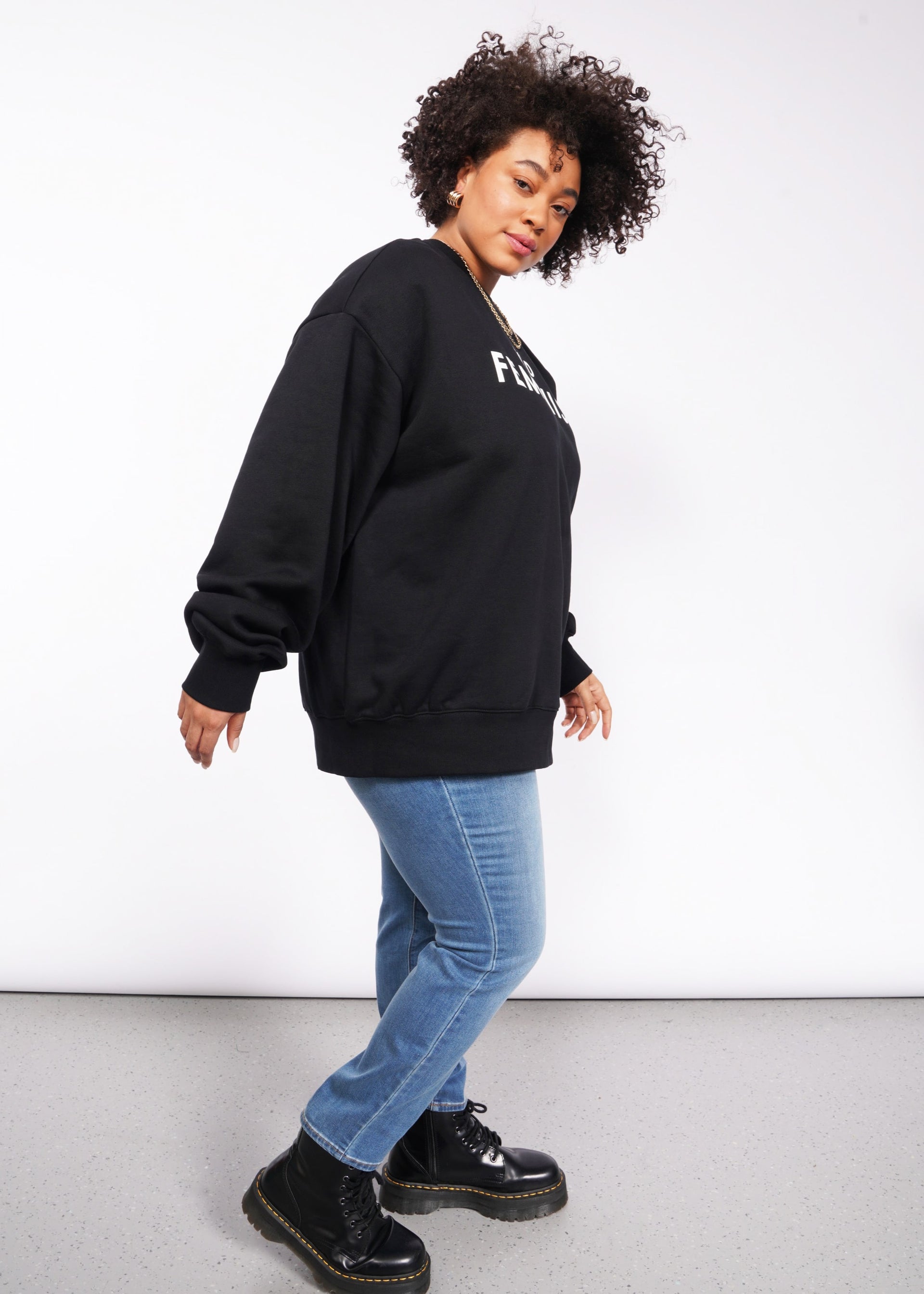 A person casually leans against a white background, wearing the Wild Feminist Fleece Crew in black with white lettering, paired with blue jeans and black boots. The brushed fleece enhances the texture of their look. They have curly hair and gaze slightly to the side.