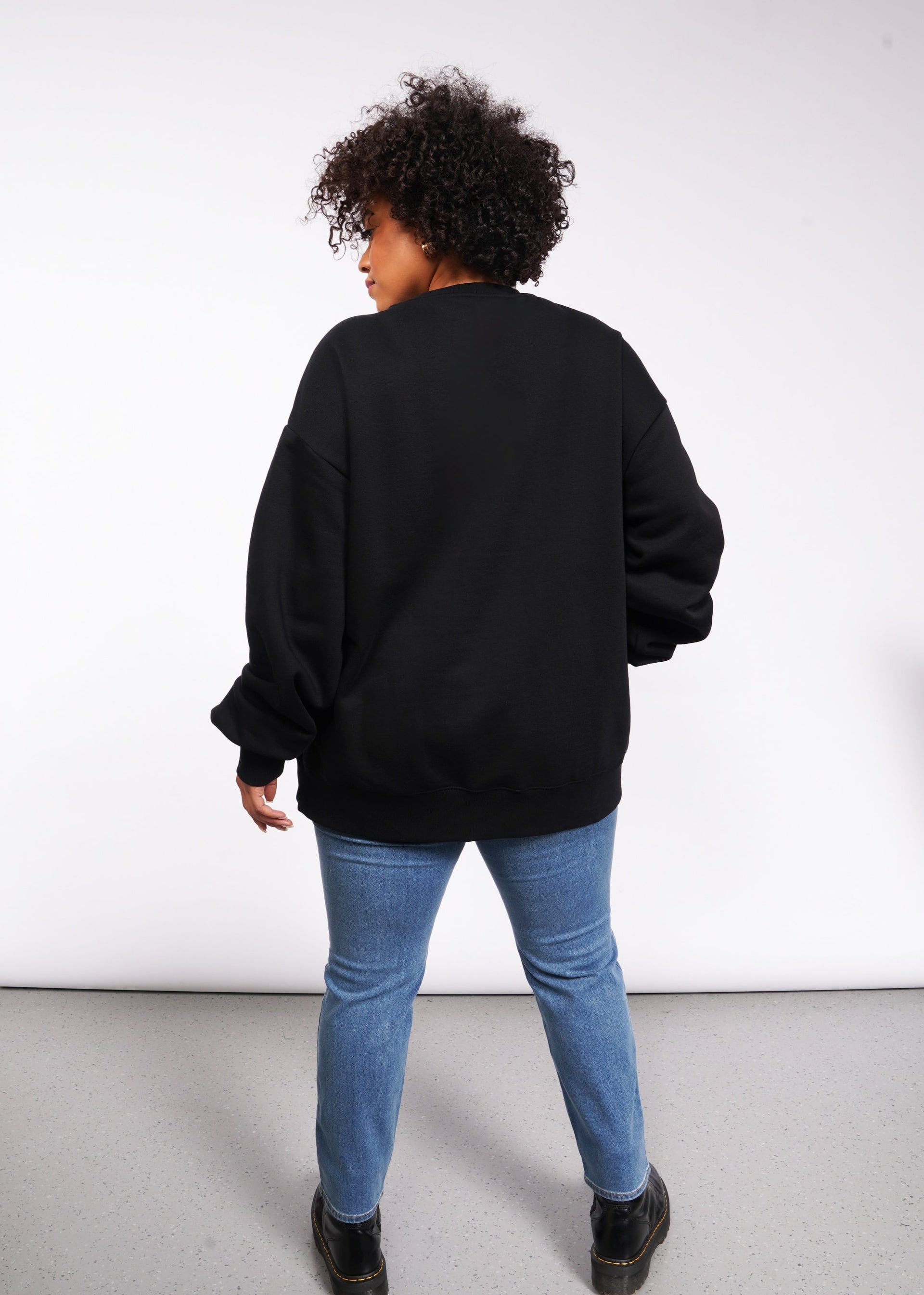 In a white room, a person with curly hair faces away wearing the Wild Feminist Fleece Crew paired with blue jeans and sturdy black boots.