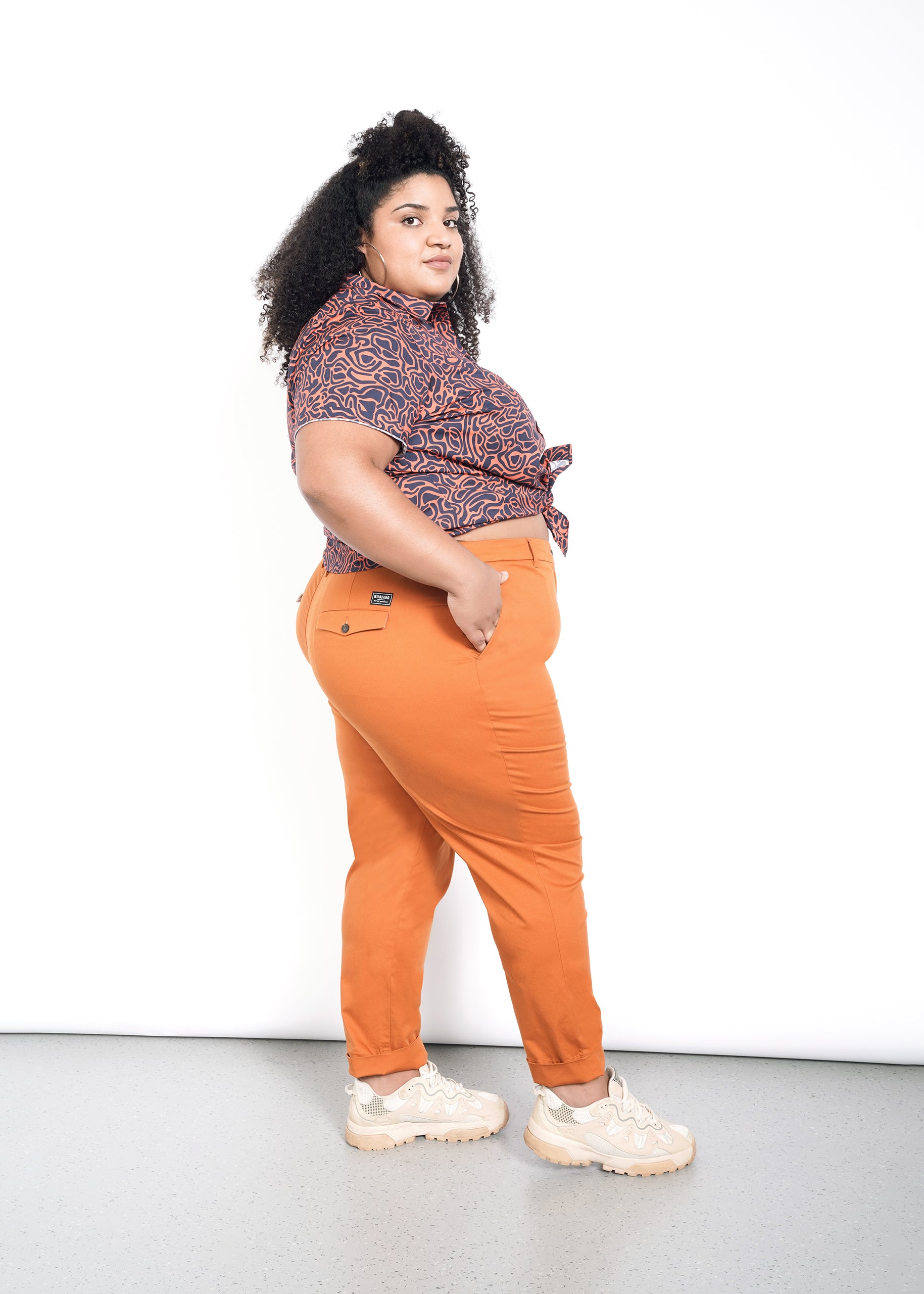 A person stands confidently against a white background, wearing a patterned short-sleeve shirt with convertible cuffs tied at the waist, The Essential Trouser in bright orange relaxed fit, and chunky beige sneakers. Their curly hair adds flair as they gaze toward the camera.